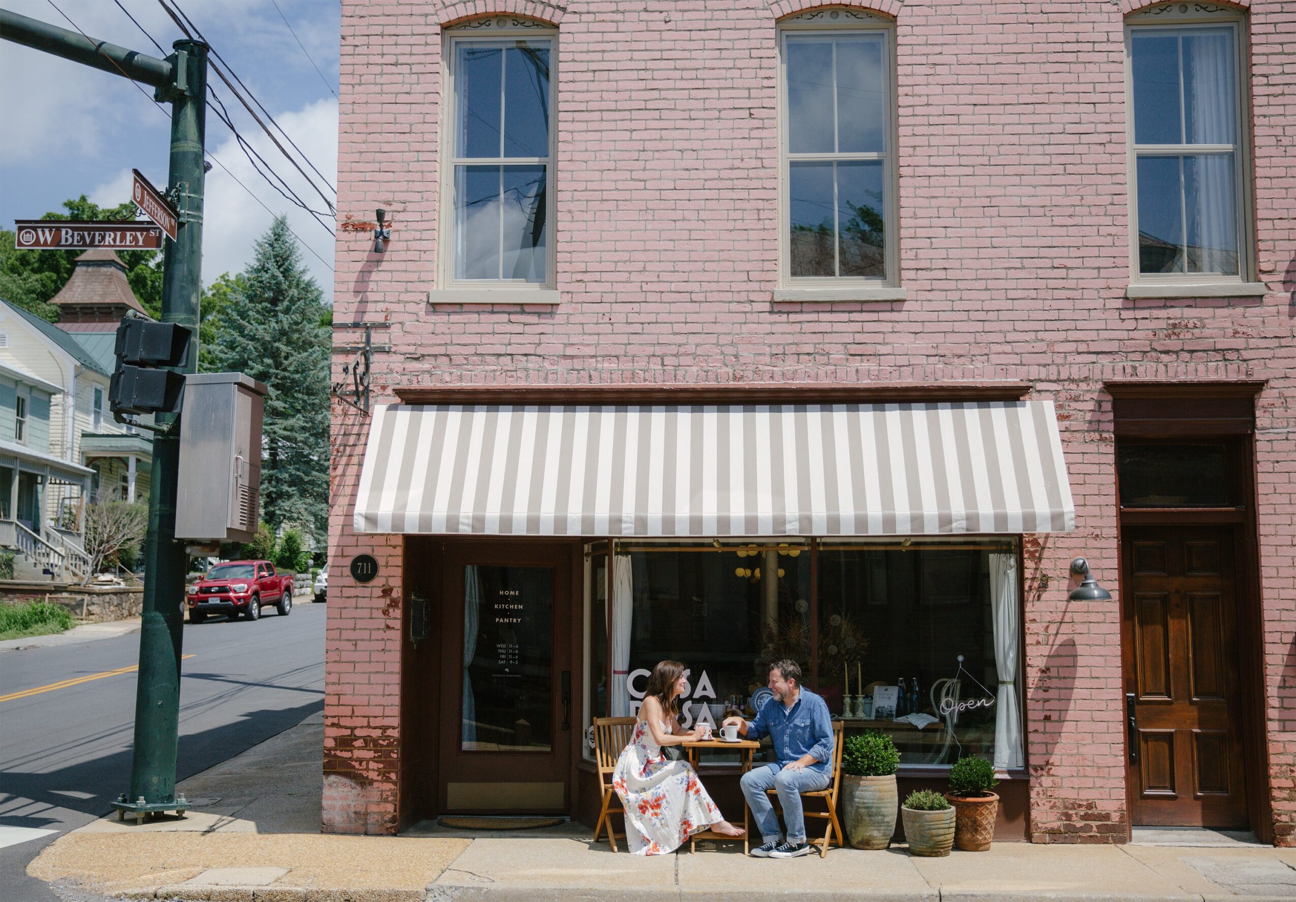 Two people sit outside a brick building with a striped awning, talking on a sunny sidewalk.