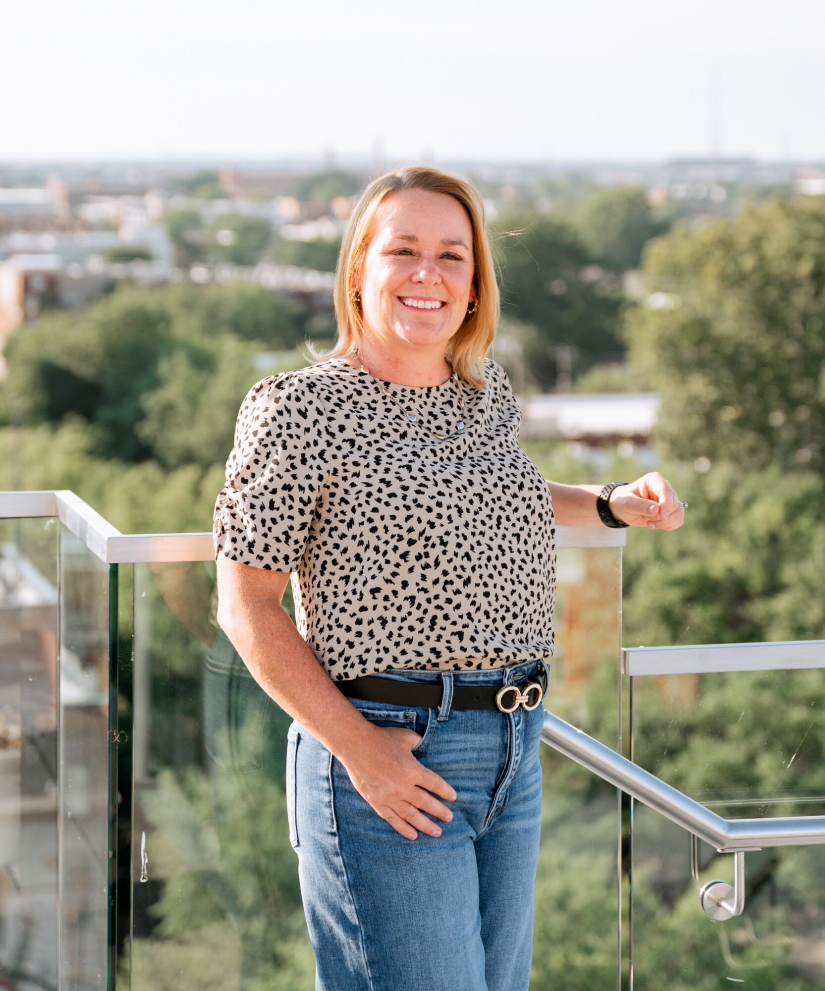 Smiling woman standing outdoors on a balcony with trees and cityscape in background.