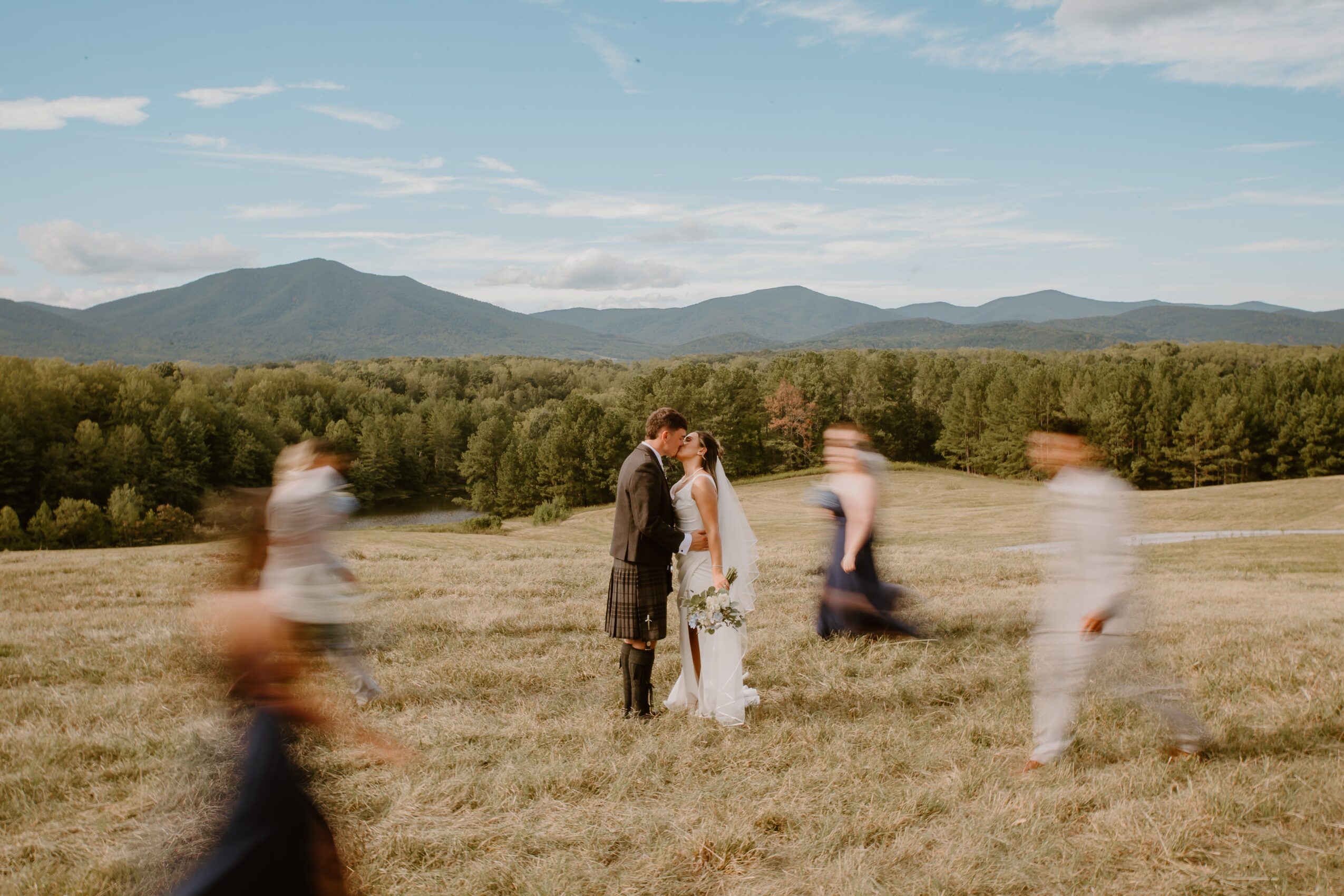 A couple stands in a field with mountains and trees in the background, surrounded by blurred people, during daytime.