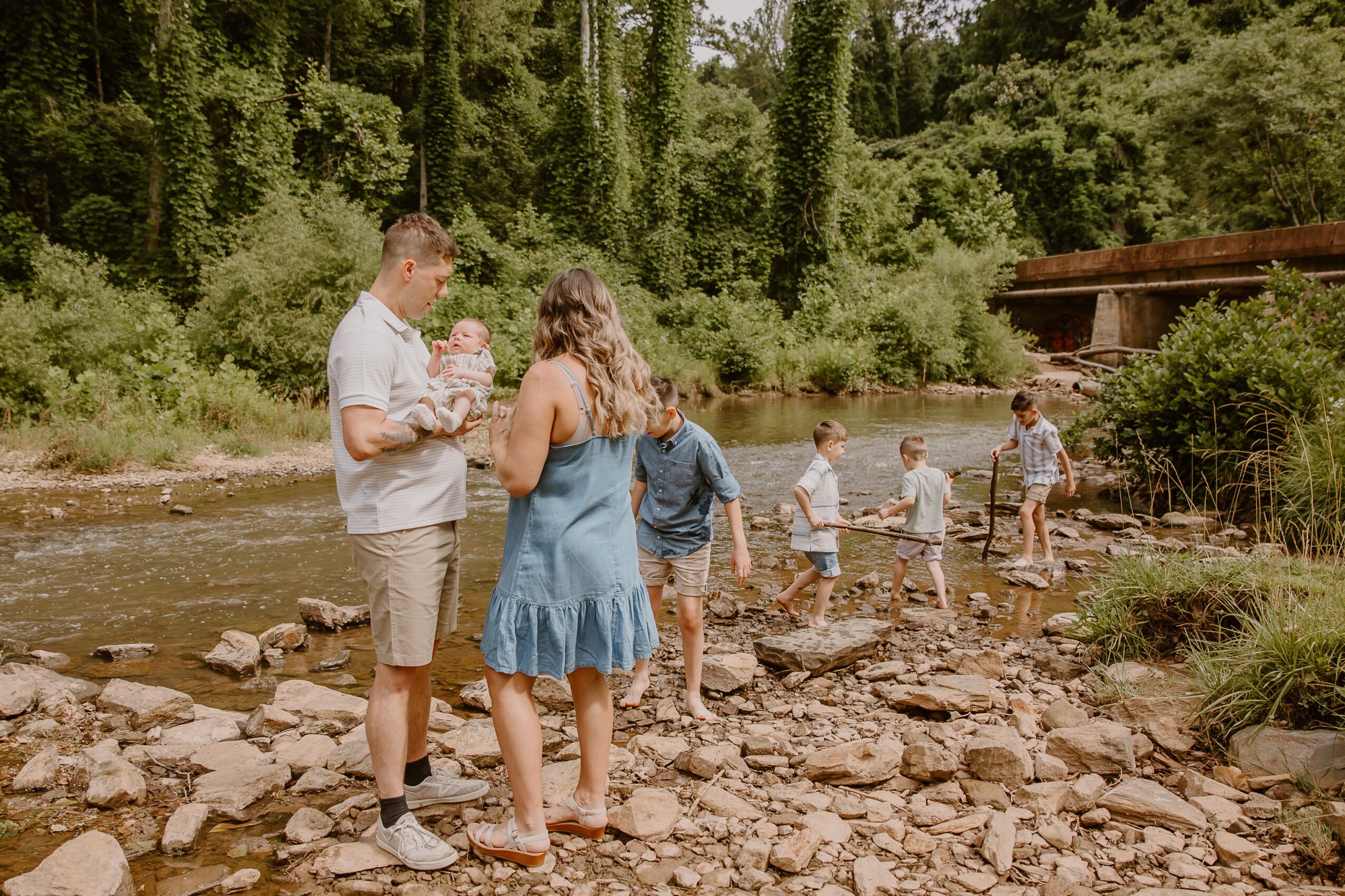 Group of people, including children and adults, by a river with trees in the background, some walking on rocks.