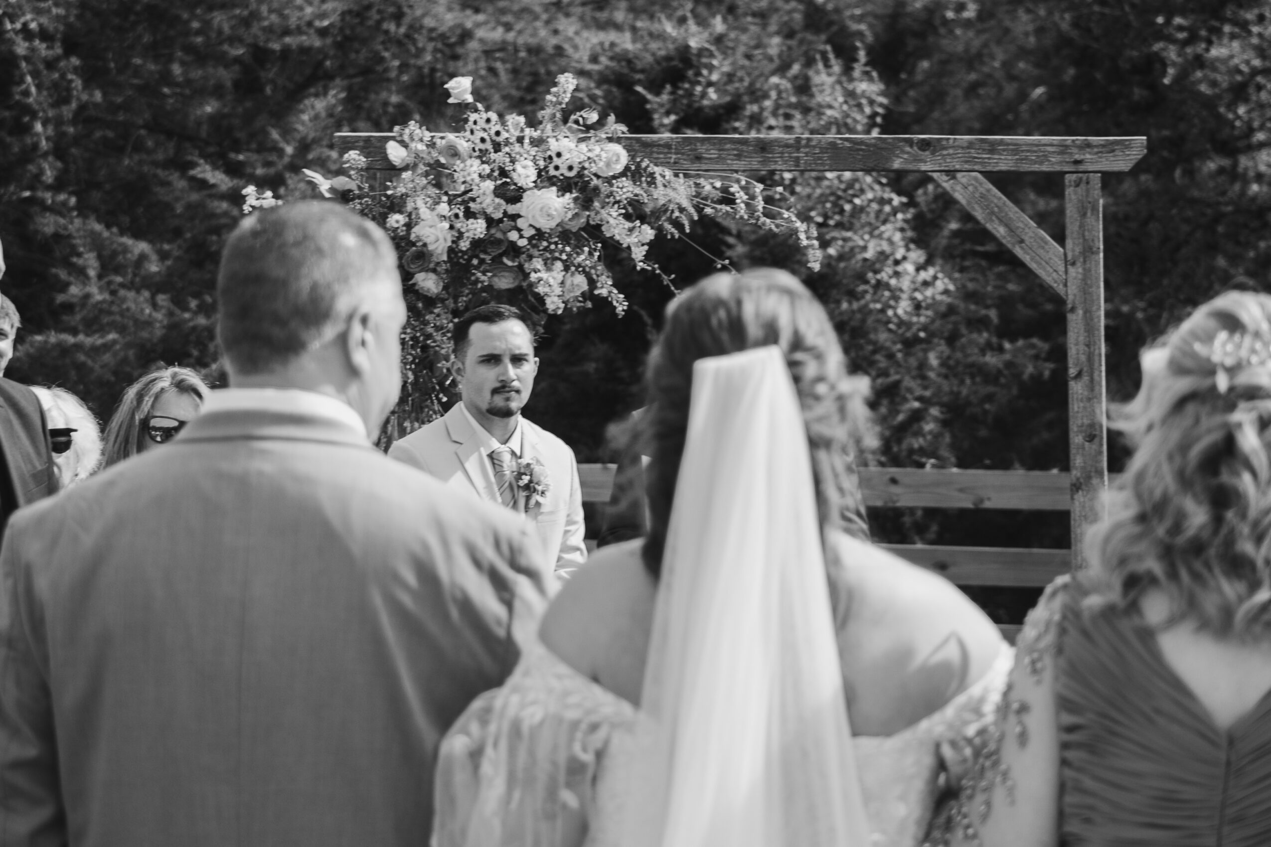 People gathered outdoors for a wedding ceremony, with a floral arch and trees in the background.