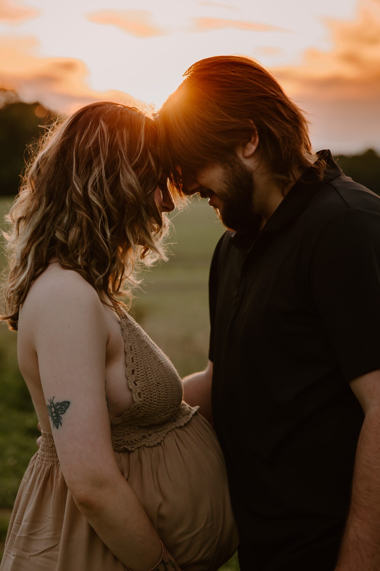 A pregnant woman and a man face each other outdoors at sunset, touching foreheads with their eyes closed.