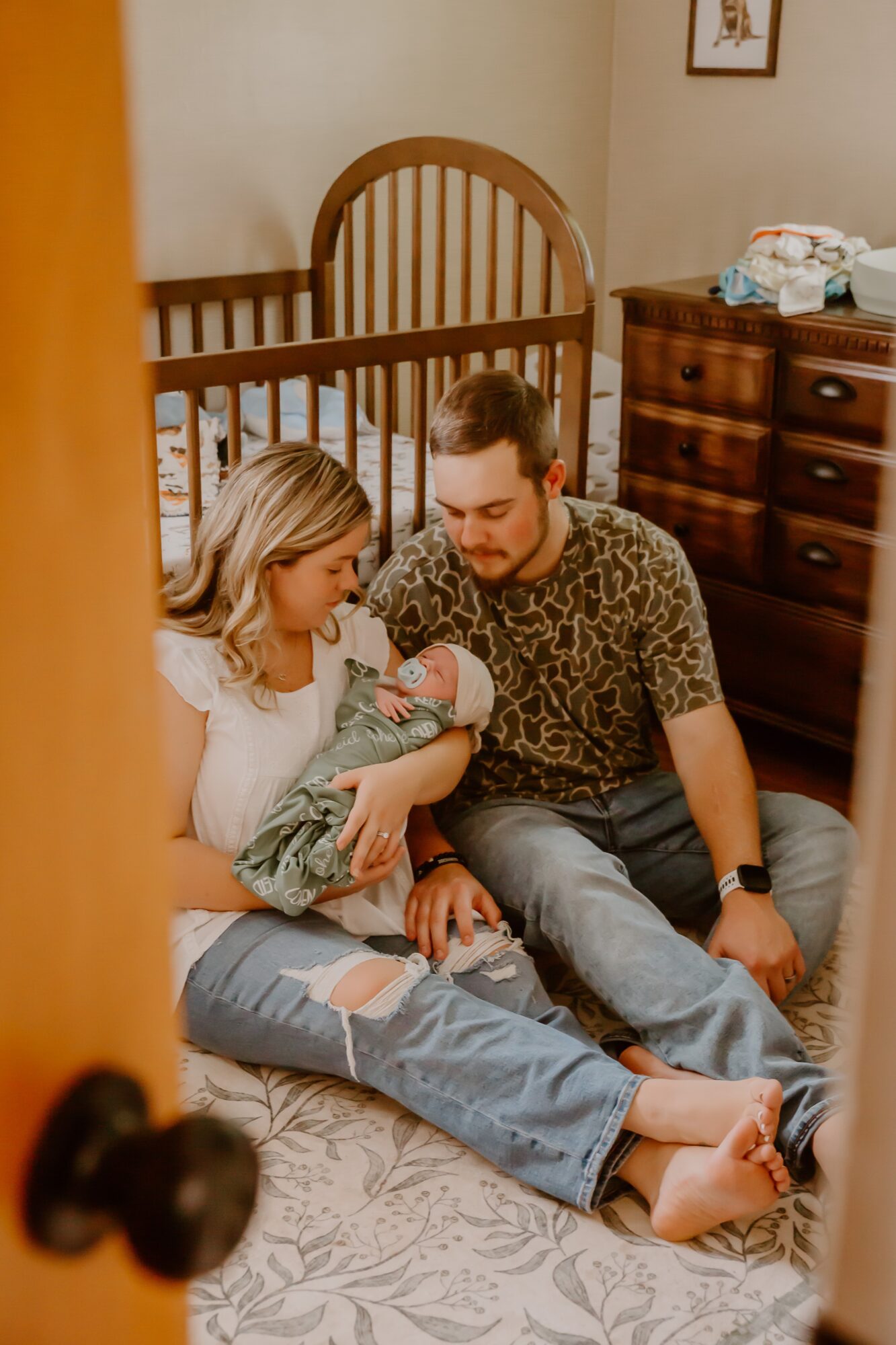 Family in nursery, woman holding newborn, man sitting beside, crib and dresser in background, warm lighting.