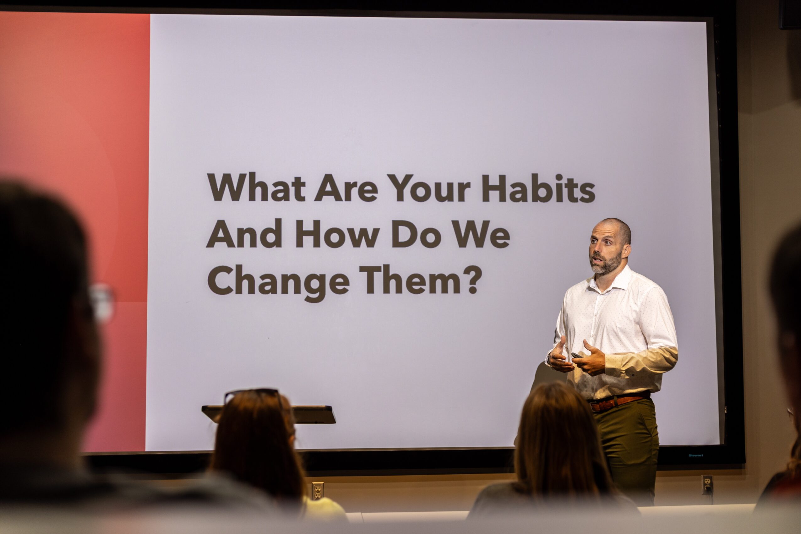 A man stands in front of a presentation slide titled 'What Are Your Habits And How Do We Change Them?' with an audience watching.
