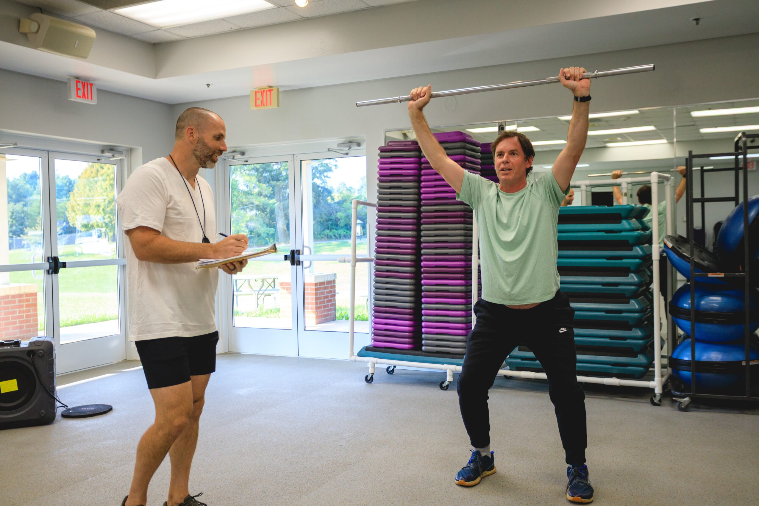 Two men in a gym, one holding a clipboard and the other lifting a barbell overhead, with exercise equipment in background.