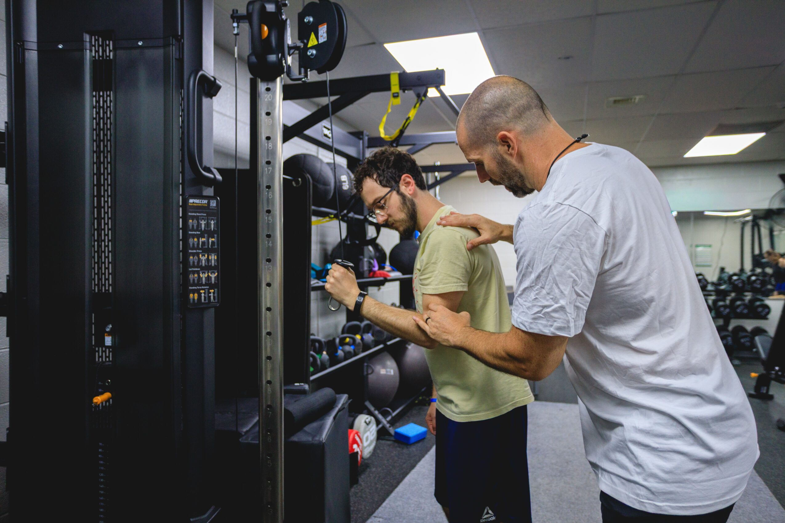 Two men working together in a gym, examining equipment, one pointing at a device, with exercise machines in background.