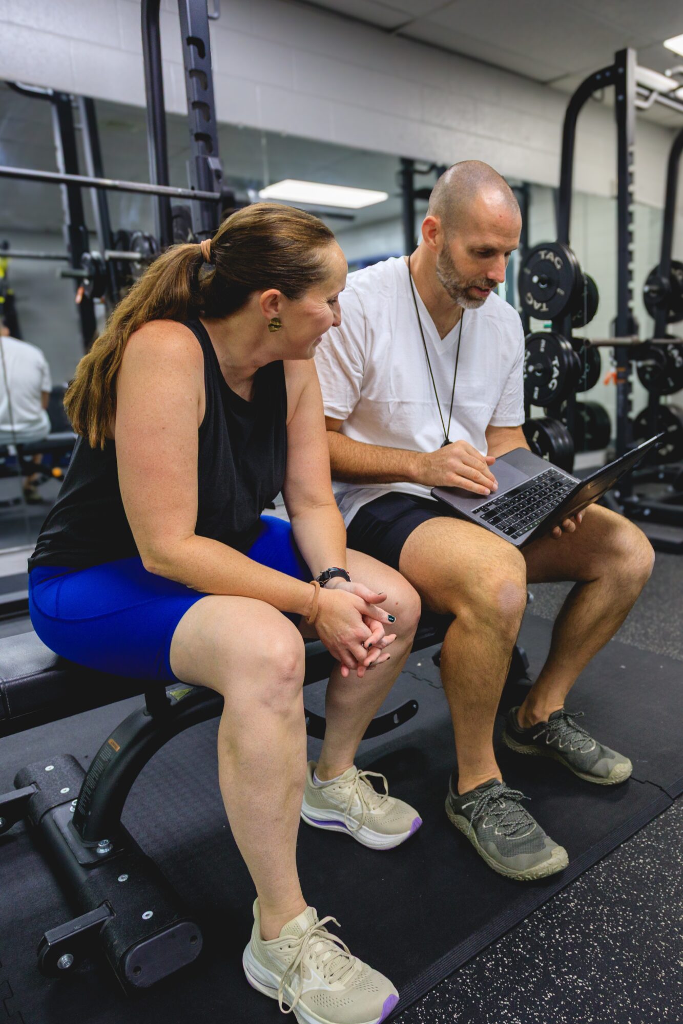 Two people sitting on a bench in a gym, one using a laptop, the other looking down, with gym equipment in background.