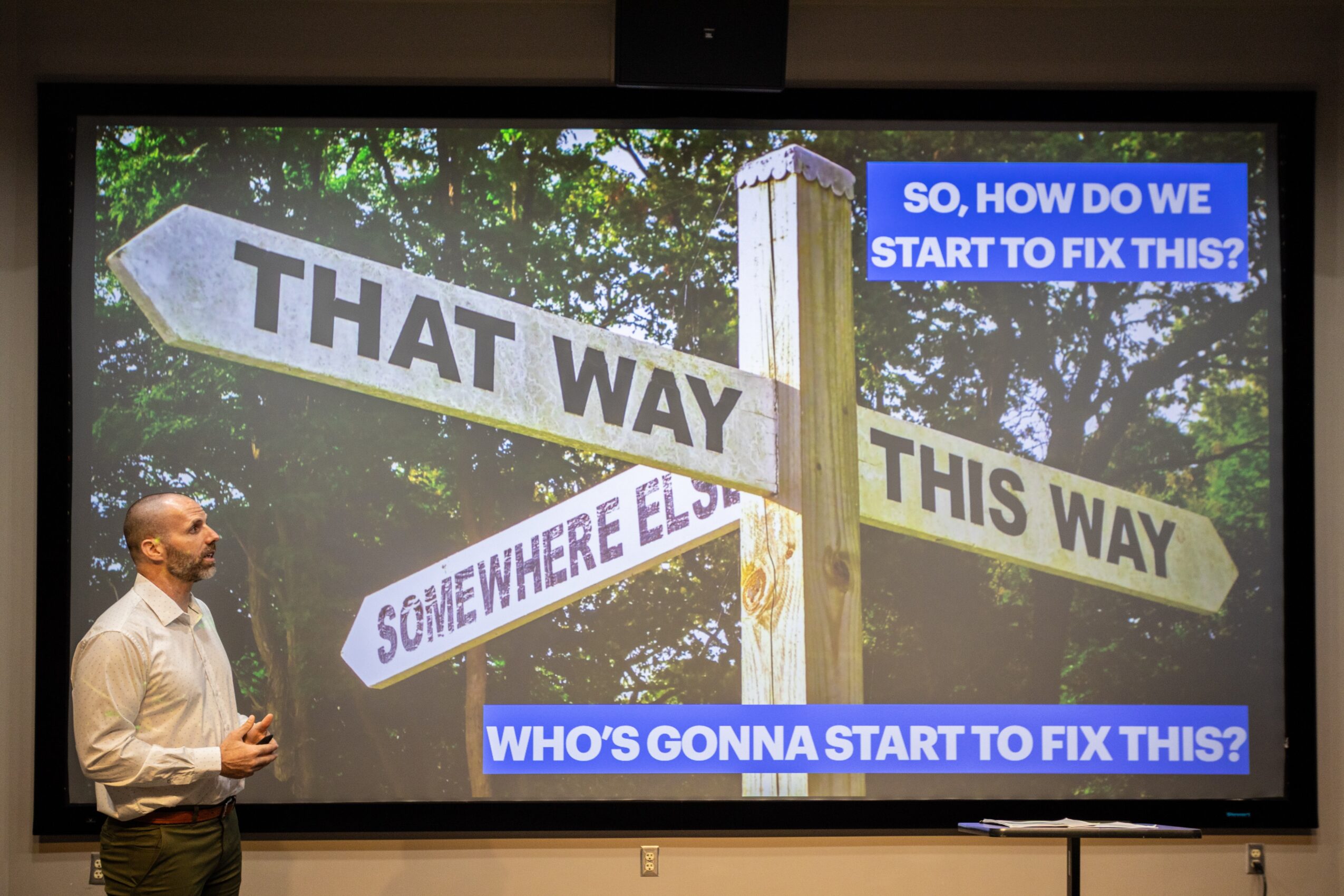 A man stands in front of a screen displaying a signpost with multiple directional signs and text overlays about fixing issues.