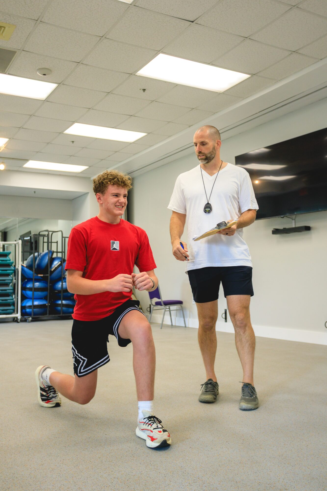 Young man in athletic clothing kneels on one knee, talking to a man holding a clipboard in a gym.