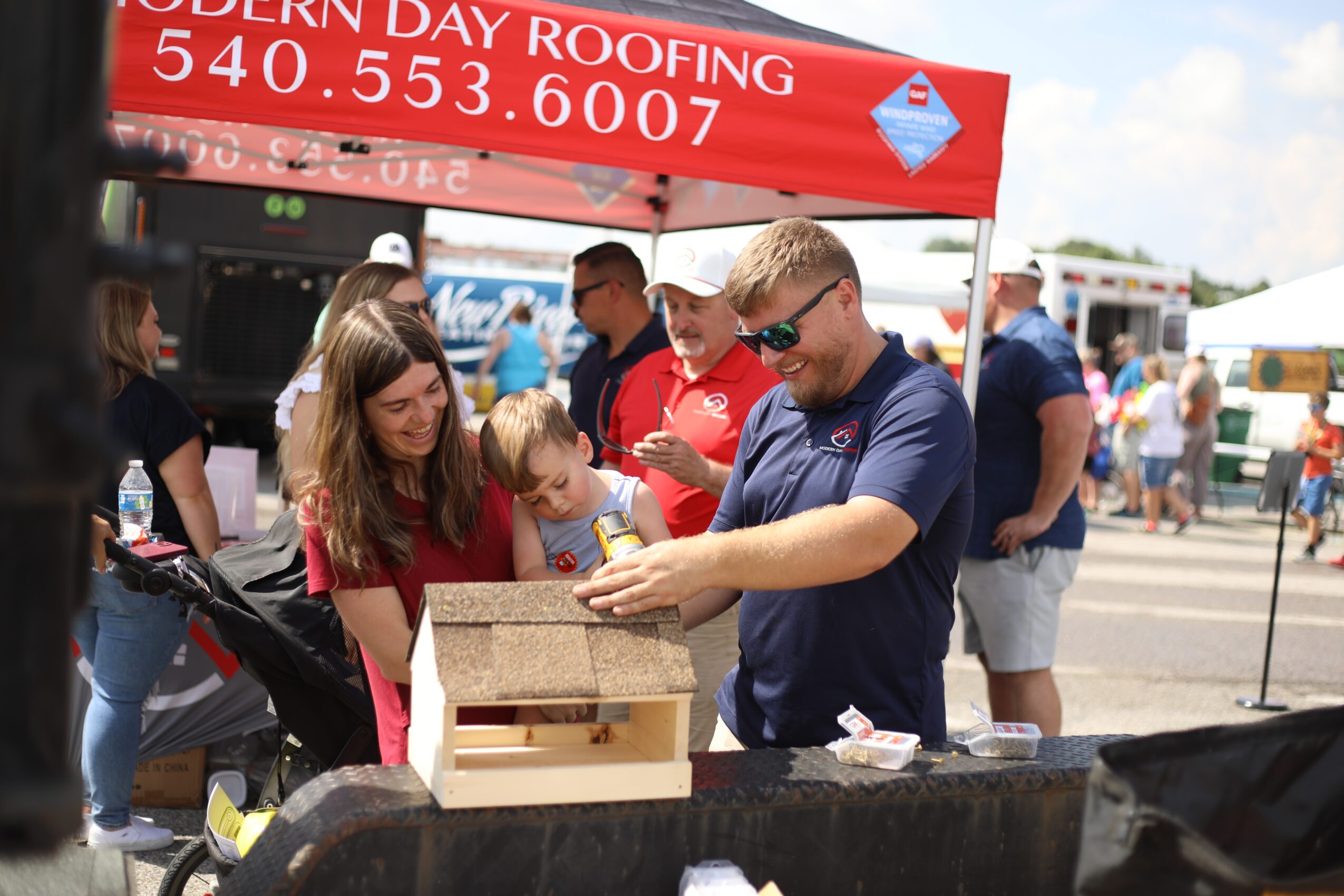 Group of people gathered around a table at an outdoor event, with a red canopy overhead, some holding drinks, and a blue sky.