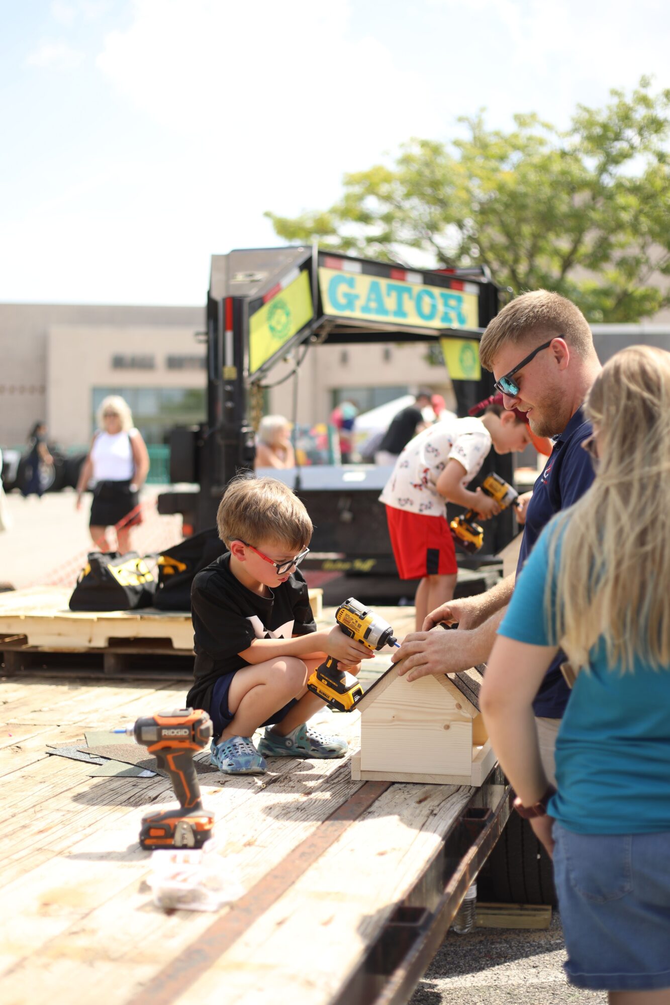 Children playing with toy tools on a wooden platform outdoors, with a person in a blue shirt in the foreground.