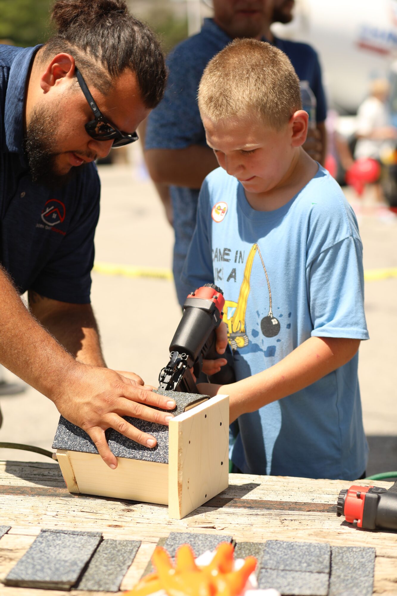 Two children and an adult working together on a small wooden project outdoors, with bricks and tools nearby.