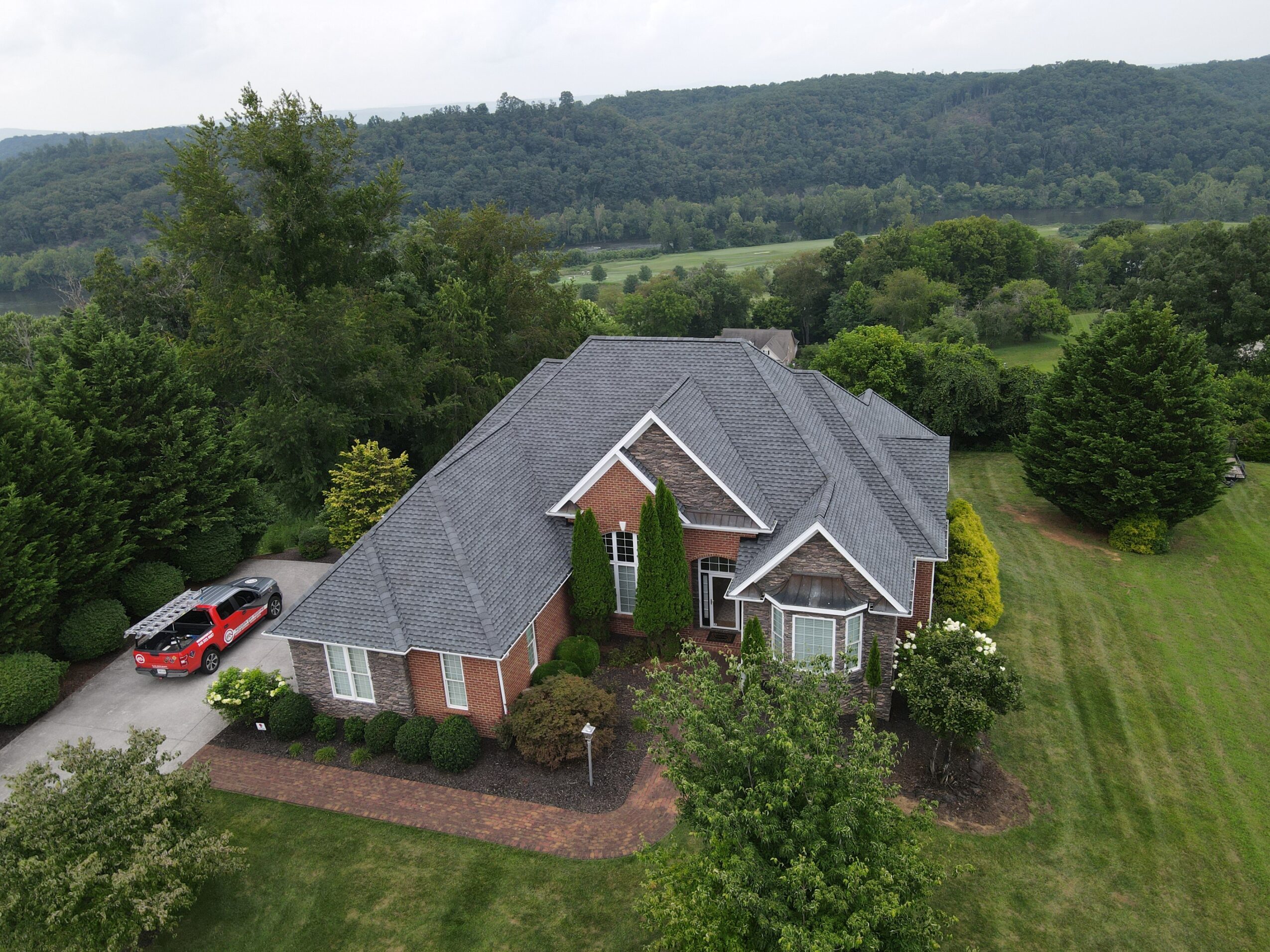 A large house with a gray roof, surrounded by trees and a lawn, with a driveway and parked car nearby.