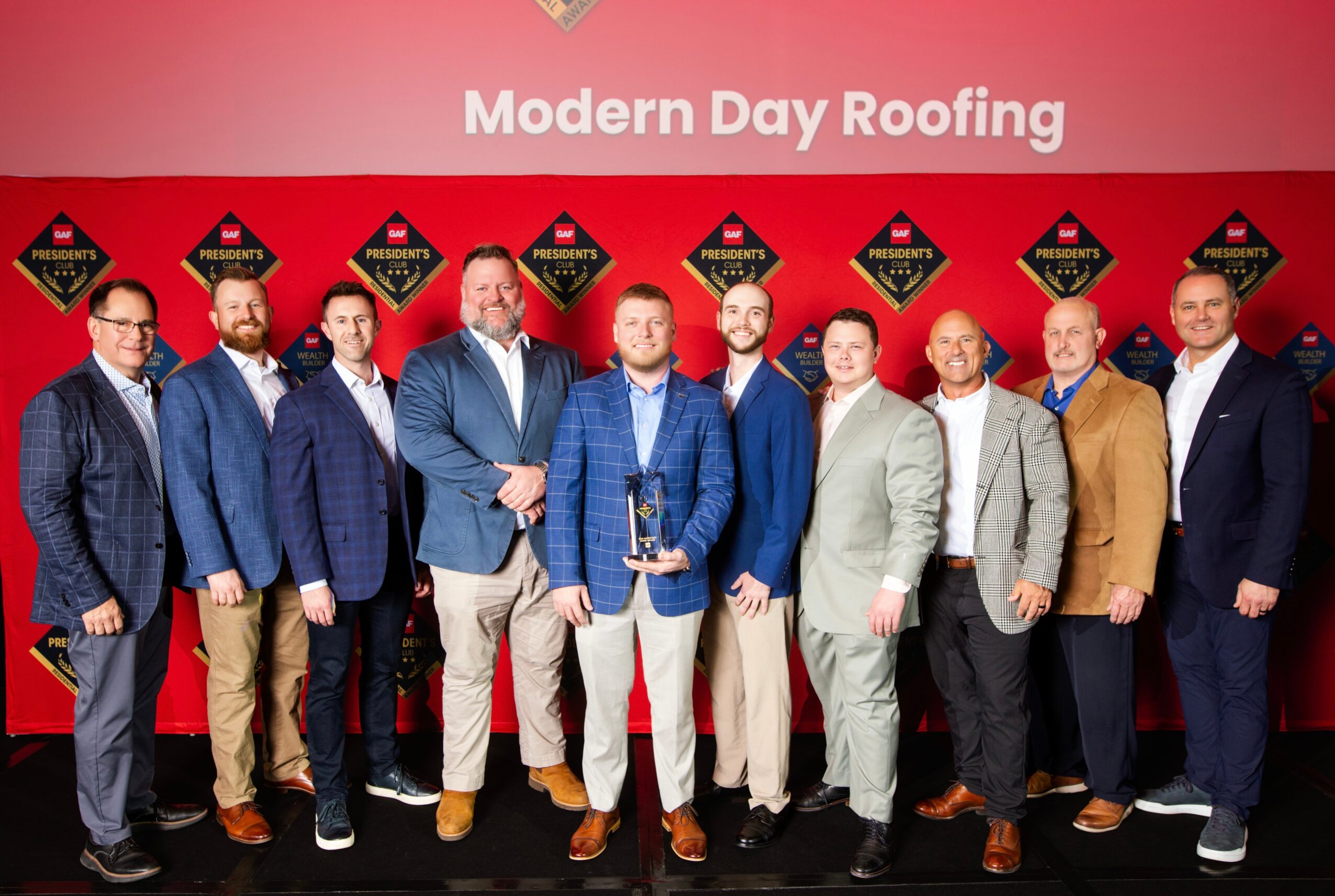 Group of ten men standing in front of a red backdrop with logos, dressed in suits and casual attire.