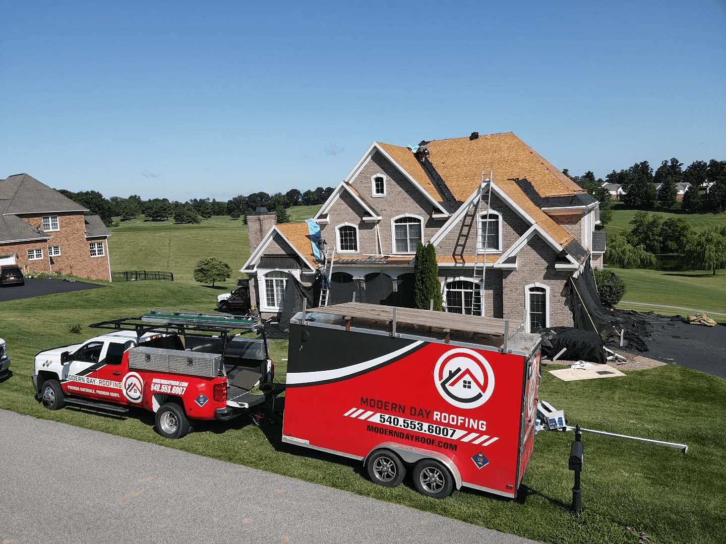 A house with a brown roof and gray walls, a red and white roofing trailer, and a red truck parked nearby.