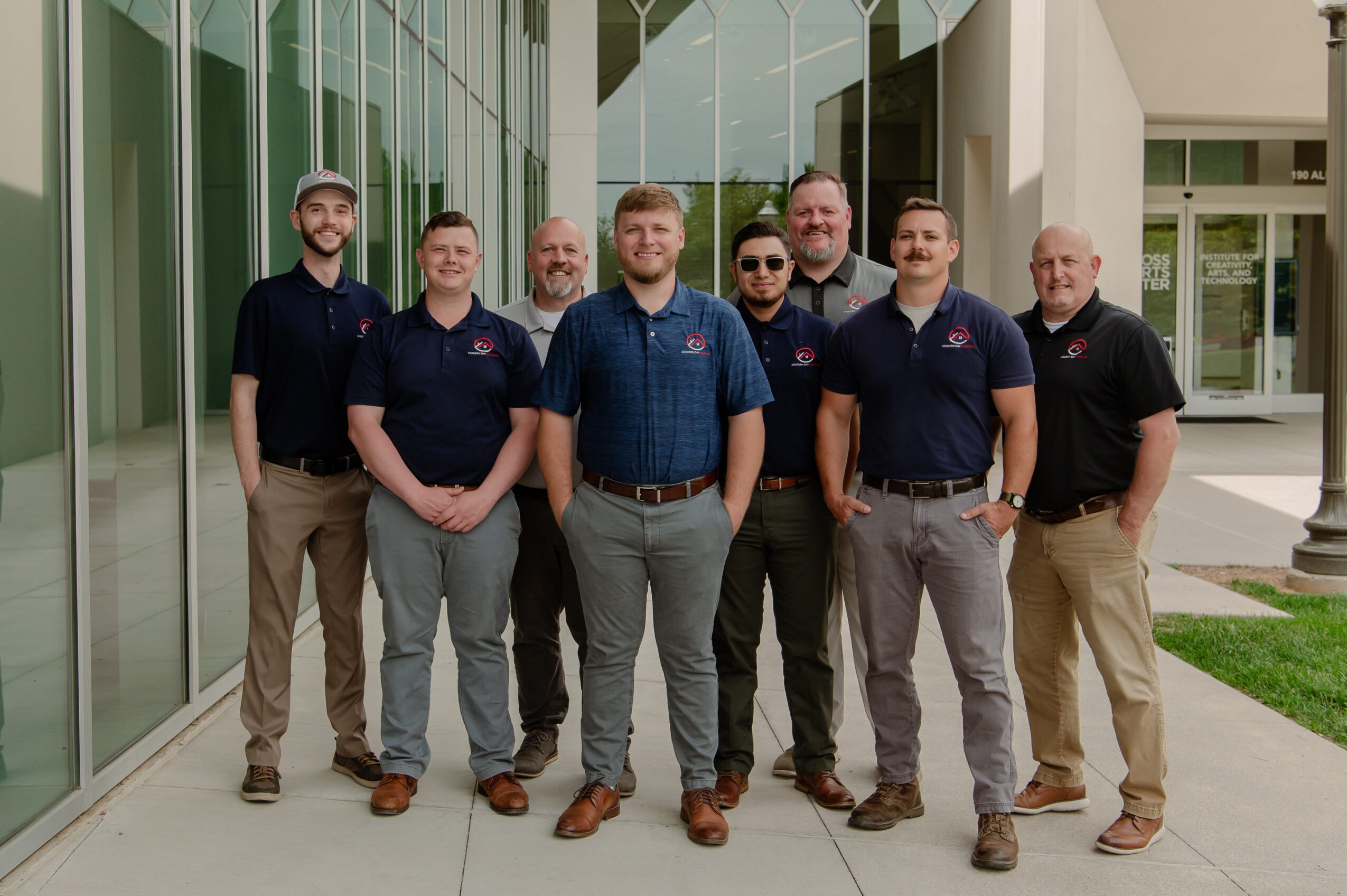 Group of seven men standing outside in front of modern glass building, smiling, wearing casual and polo shirts.
