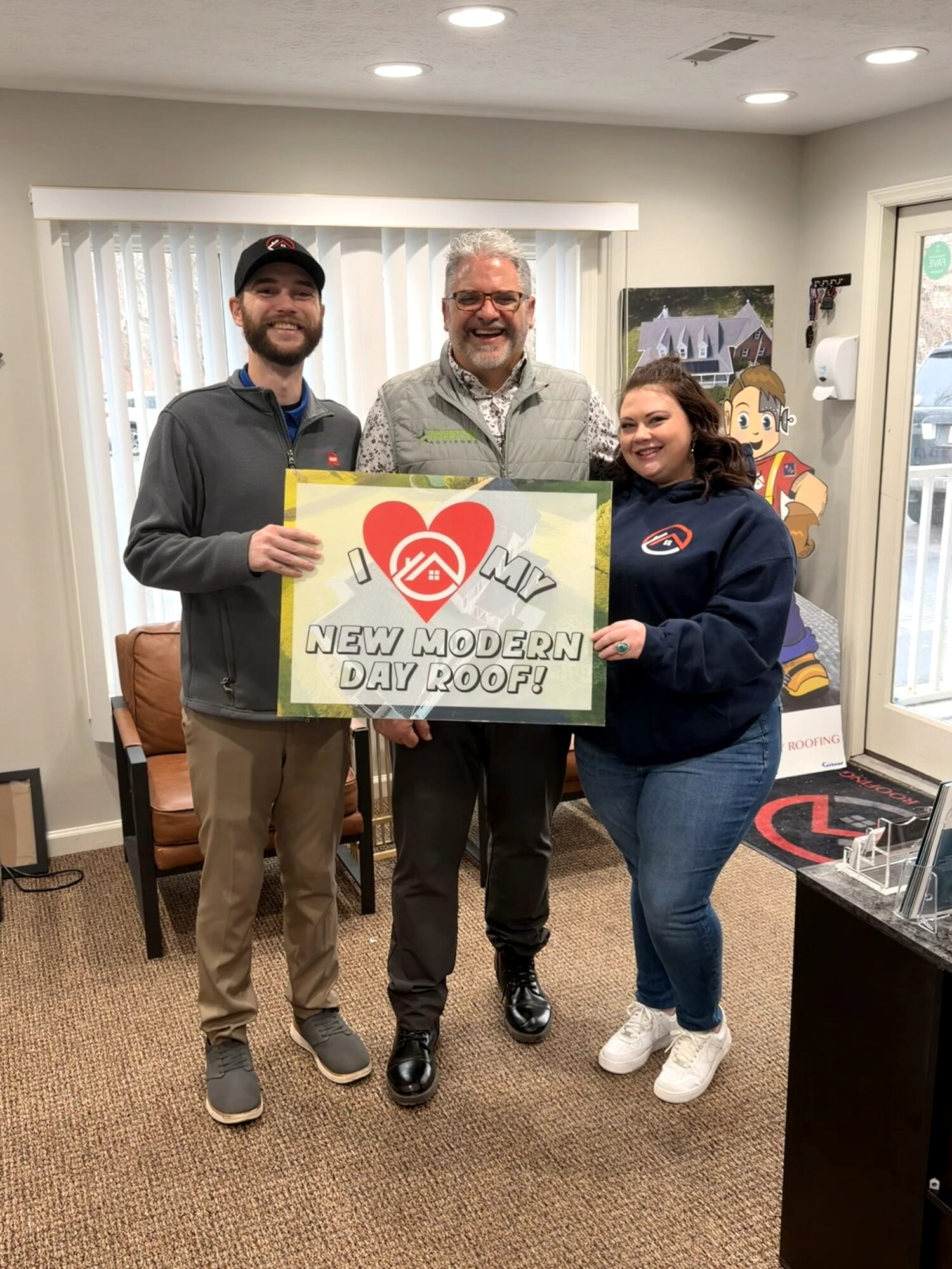 Three people standing indoors holding a sign that reads 'New Modern Day Roof!' with a heart logo, smiling at the camera.