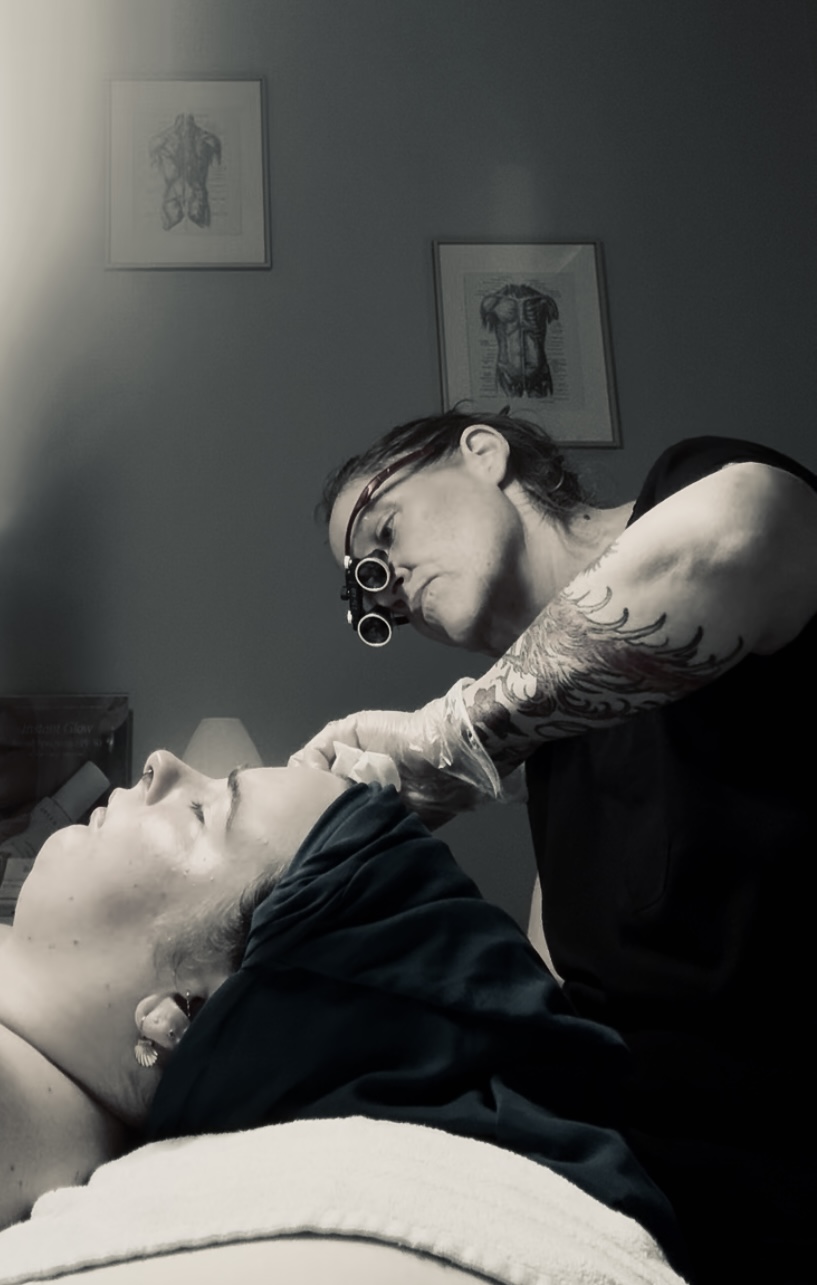 Tattooed person wearing glasses leans over a person lying down, in a room with framed artwork on the wall.
