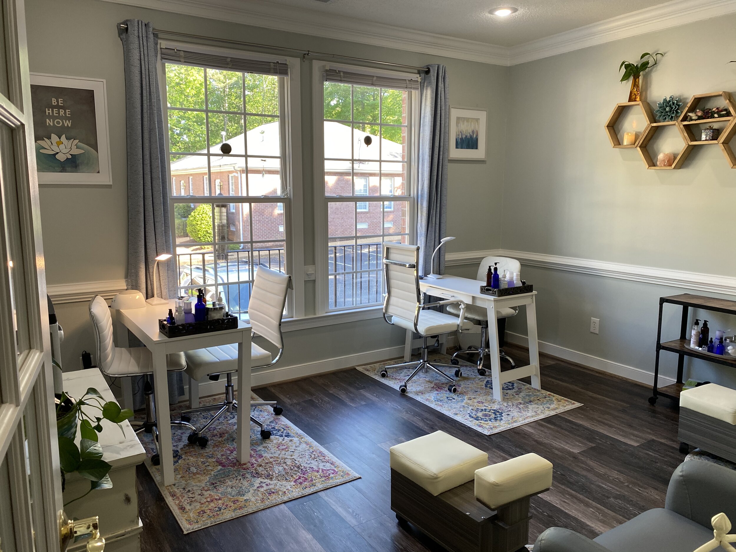 Bright room with two desks near large windows, decorated with plants, wall art, and hexagonal shelves, with a dark wood floor.