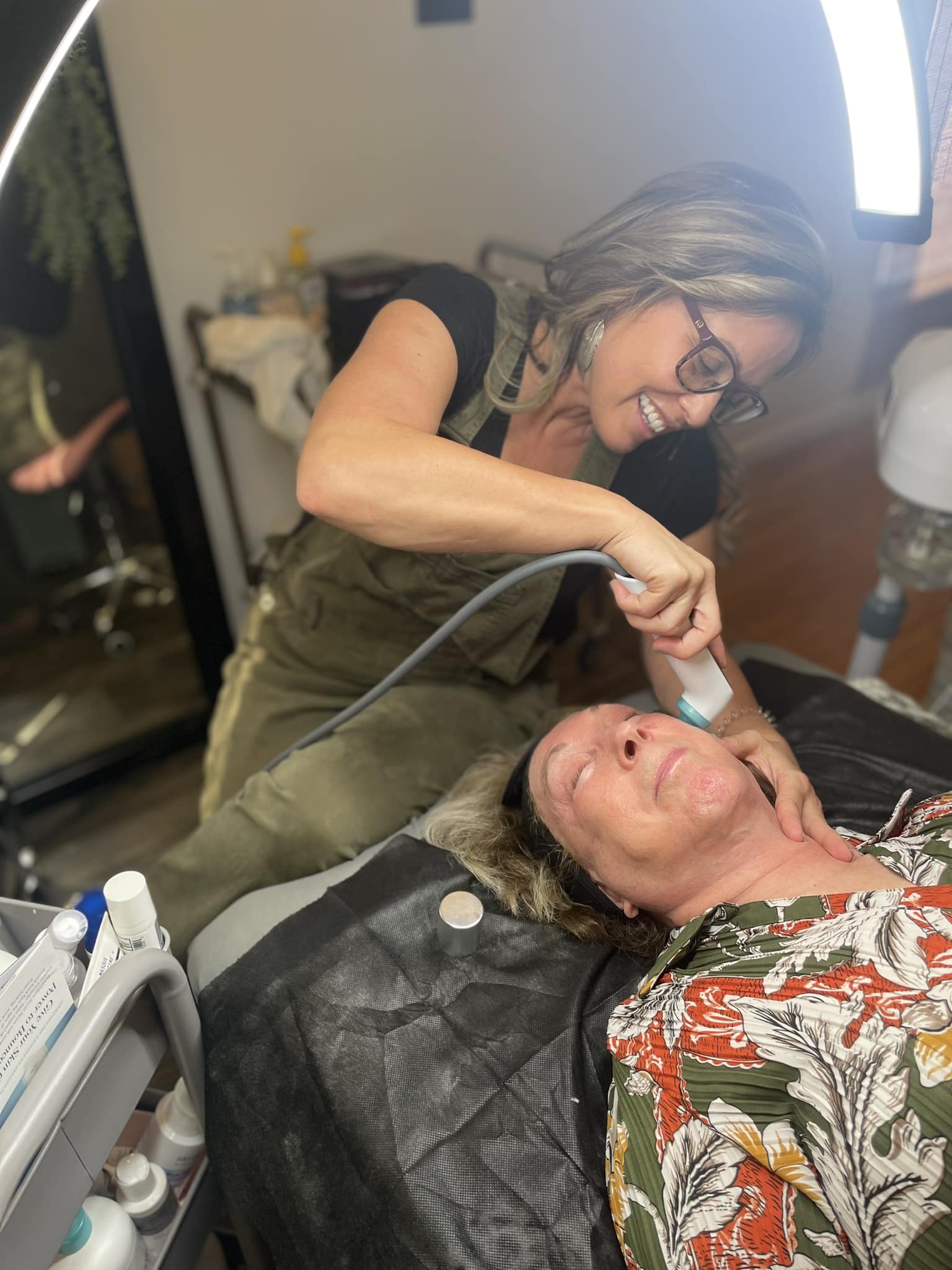Woman with glasses and curly hair performs a procedure on a woman lying down, in a clinical setting.
