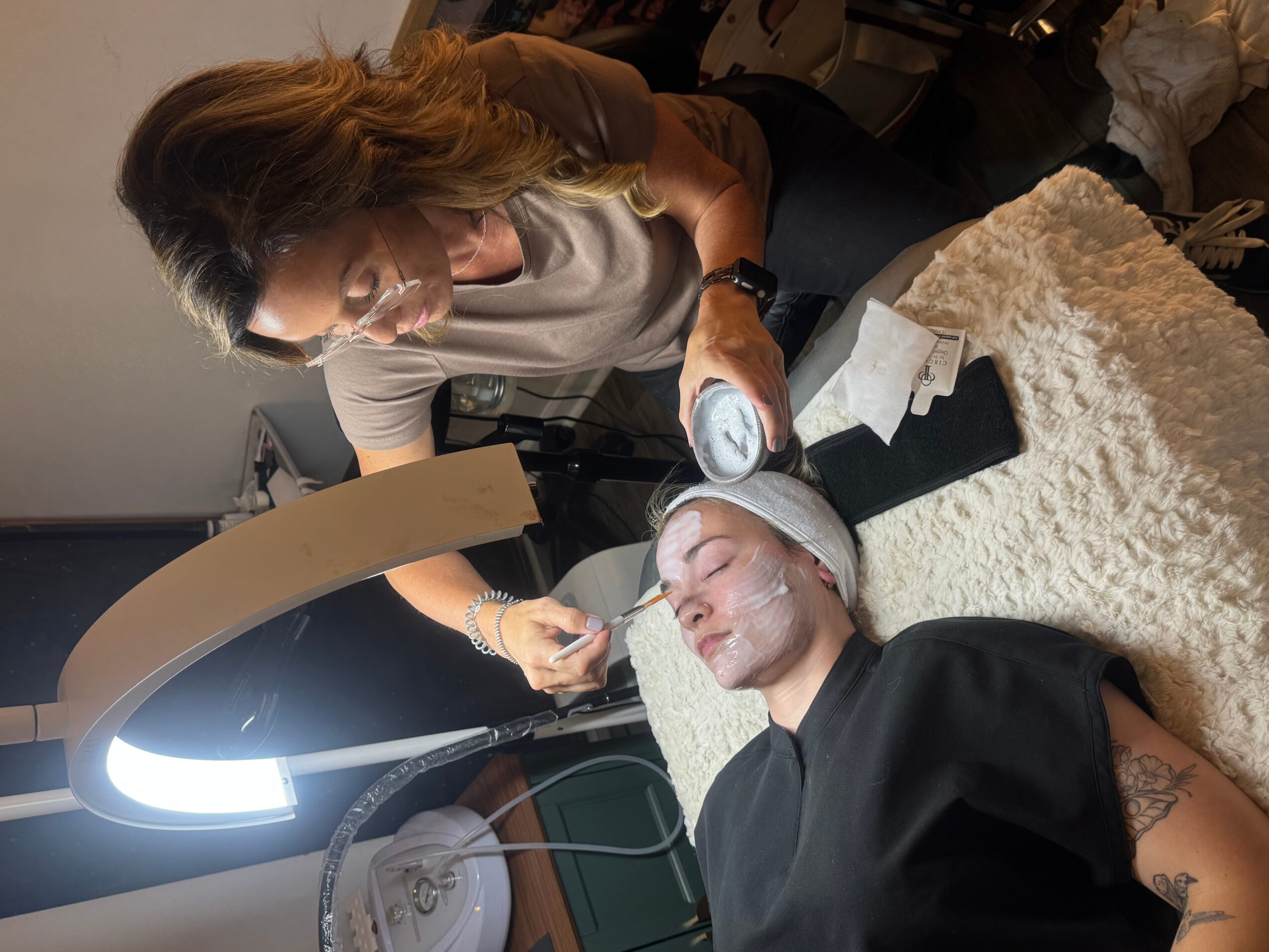 A woman performs a facial treatment on a person lying down with eyes closed, in a clinical setting.