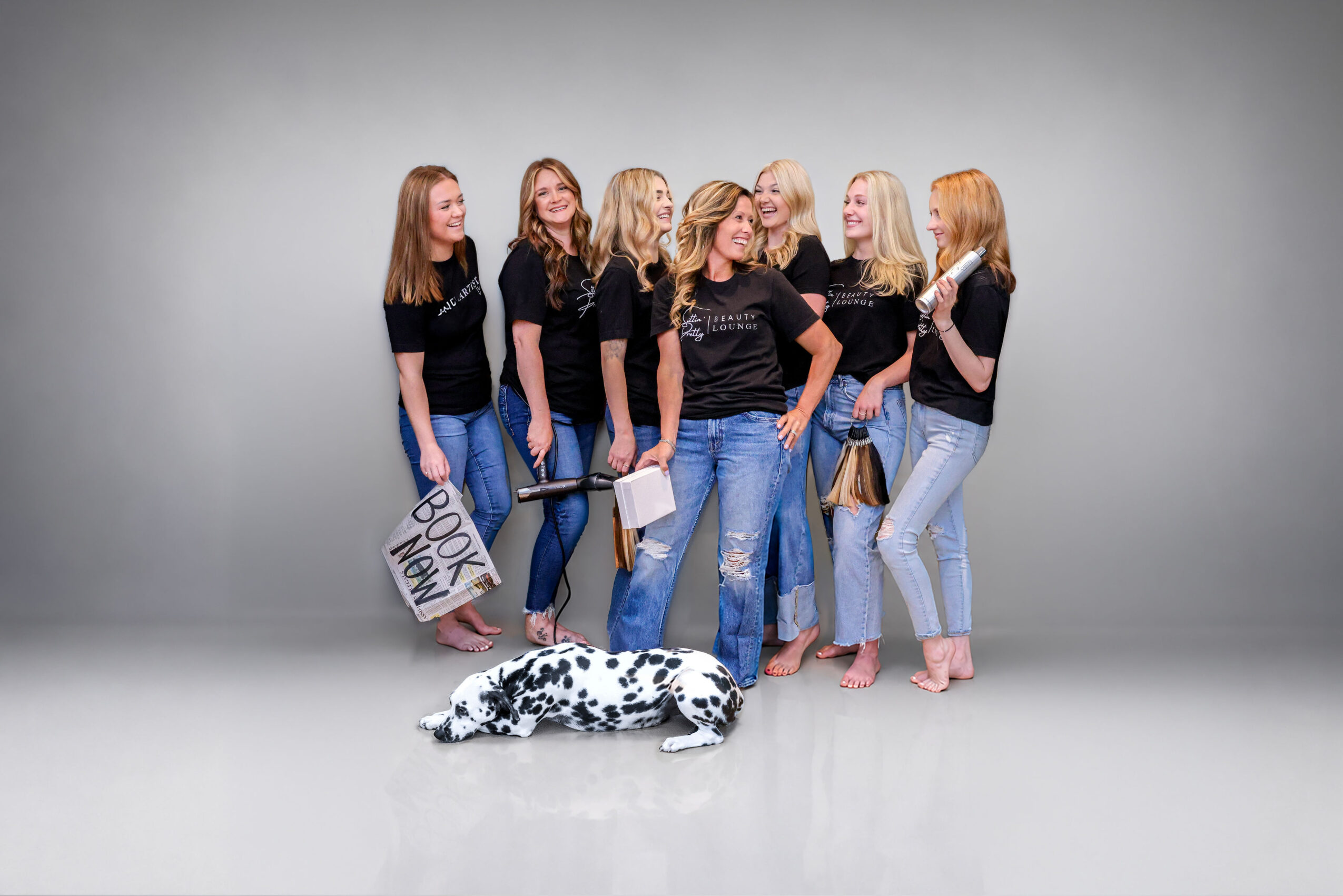 Seven women stand together in a studio, with a Dalmatian dog lying on the floor in front of them, all facing each other.