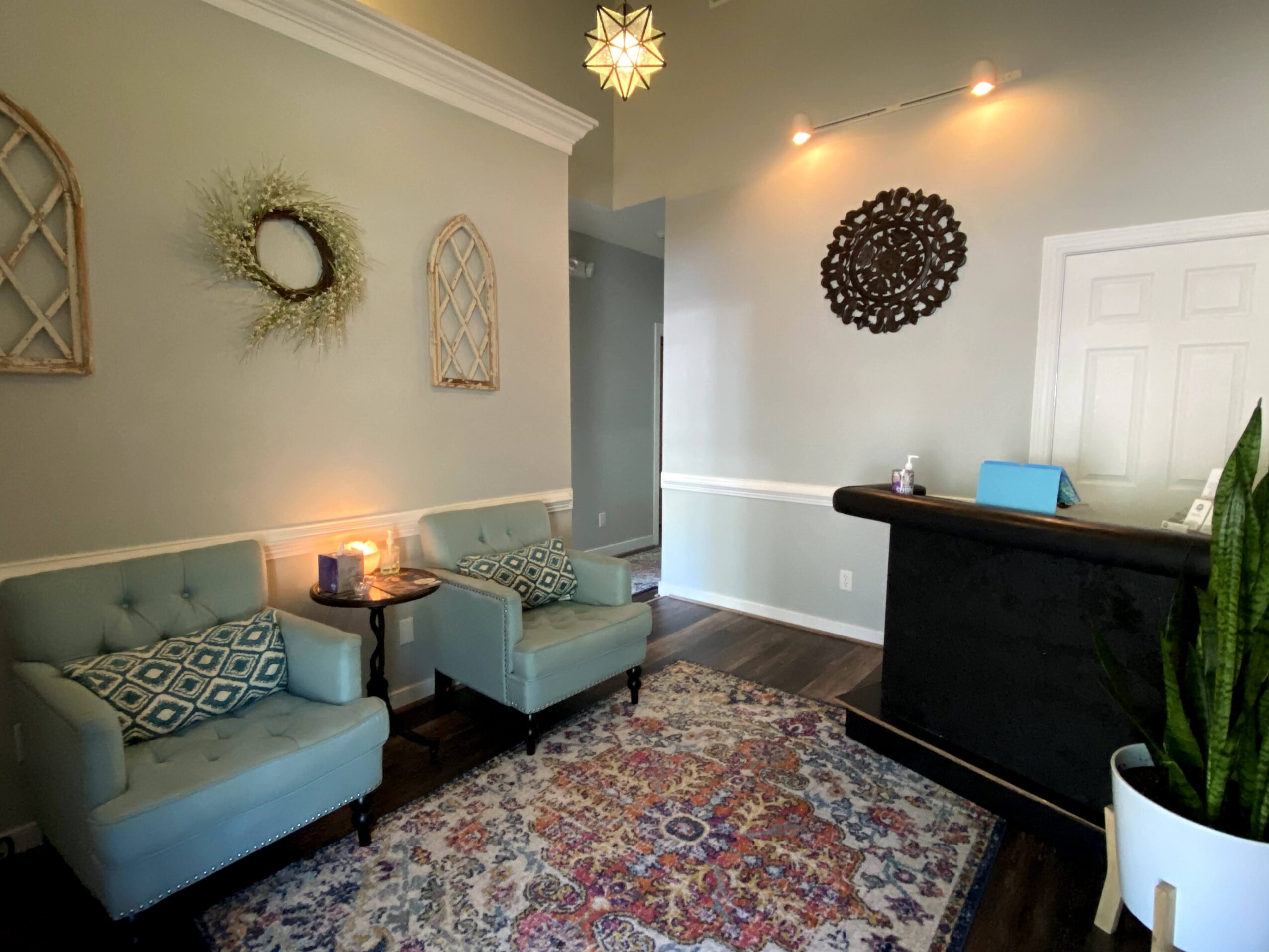 Living room with two light blue armchairs, a patterned rug, a dark reception desk, and wall decorations, including a wreath and a round wall art.