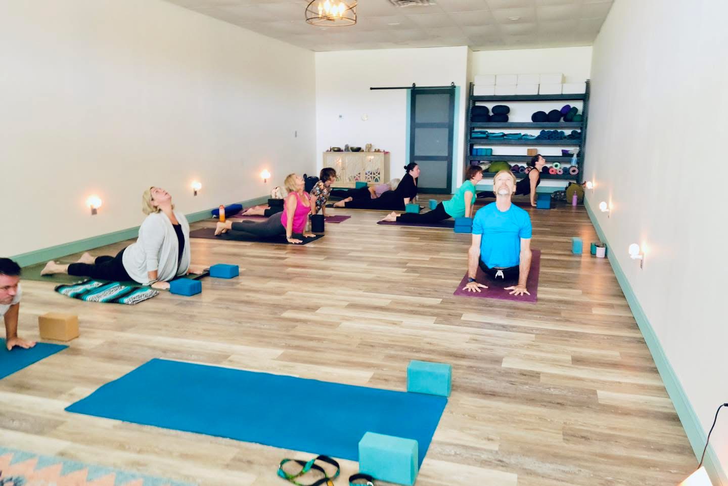 People practicing yoga in a spacious room with wooden floors, mats, and shelves with supplies, some sitting and some in poses.