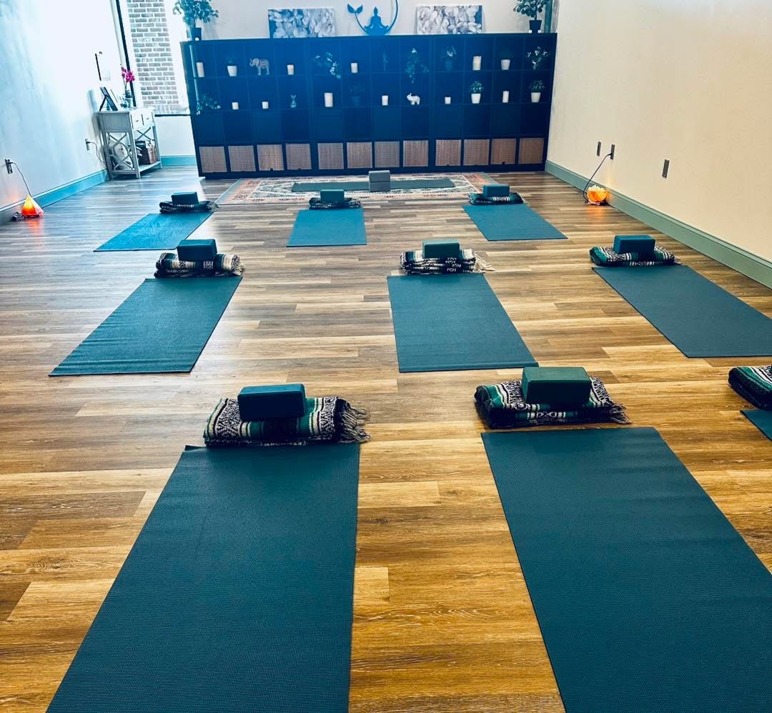Yoga mats with rolled towels and blocks arranged in a spacious room with wooden flooring and a large window.