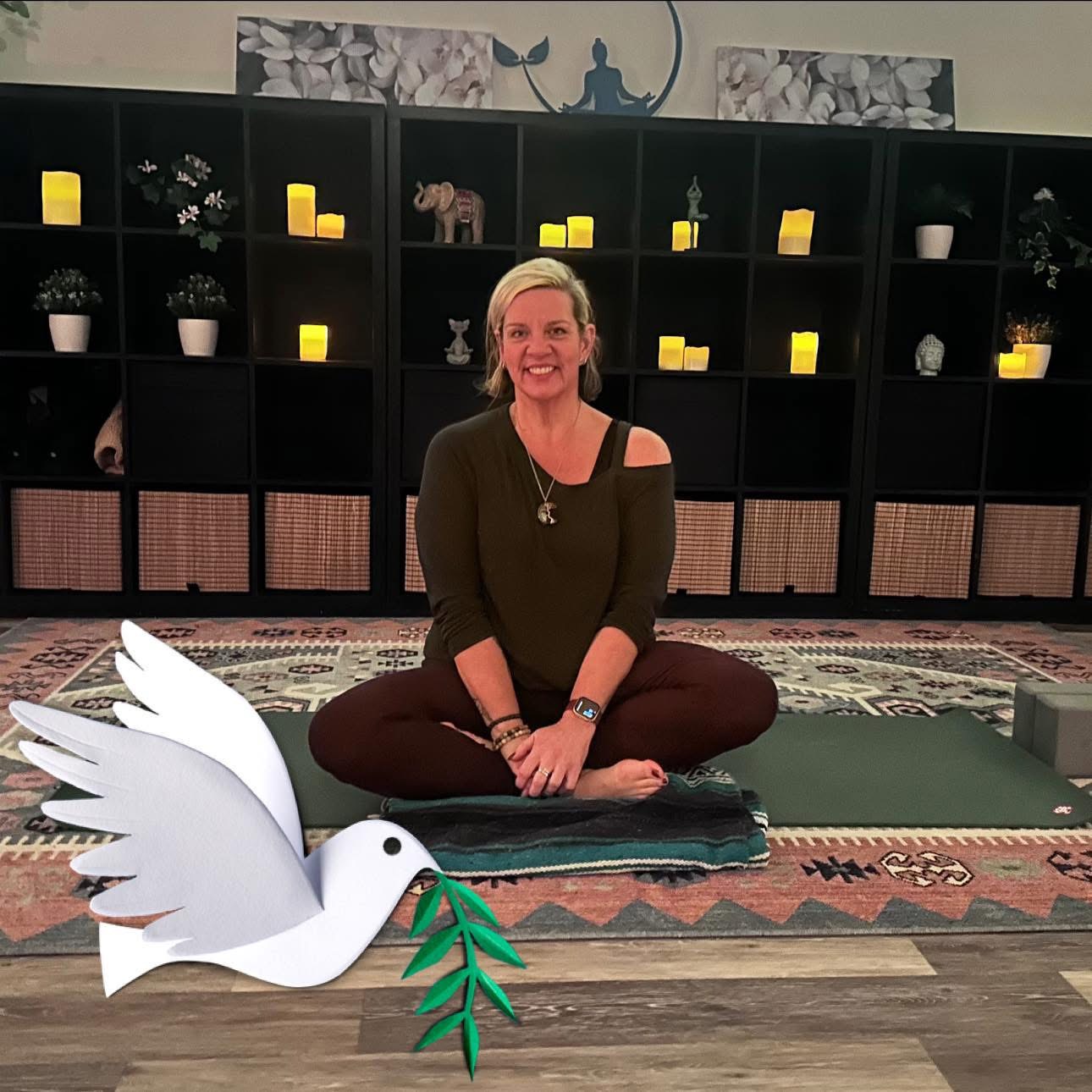 Woman sitting cross-legged on a mat indoors, smiling, with shelves and candles in the background.