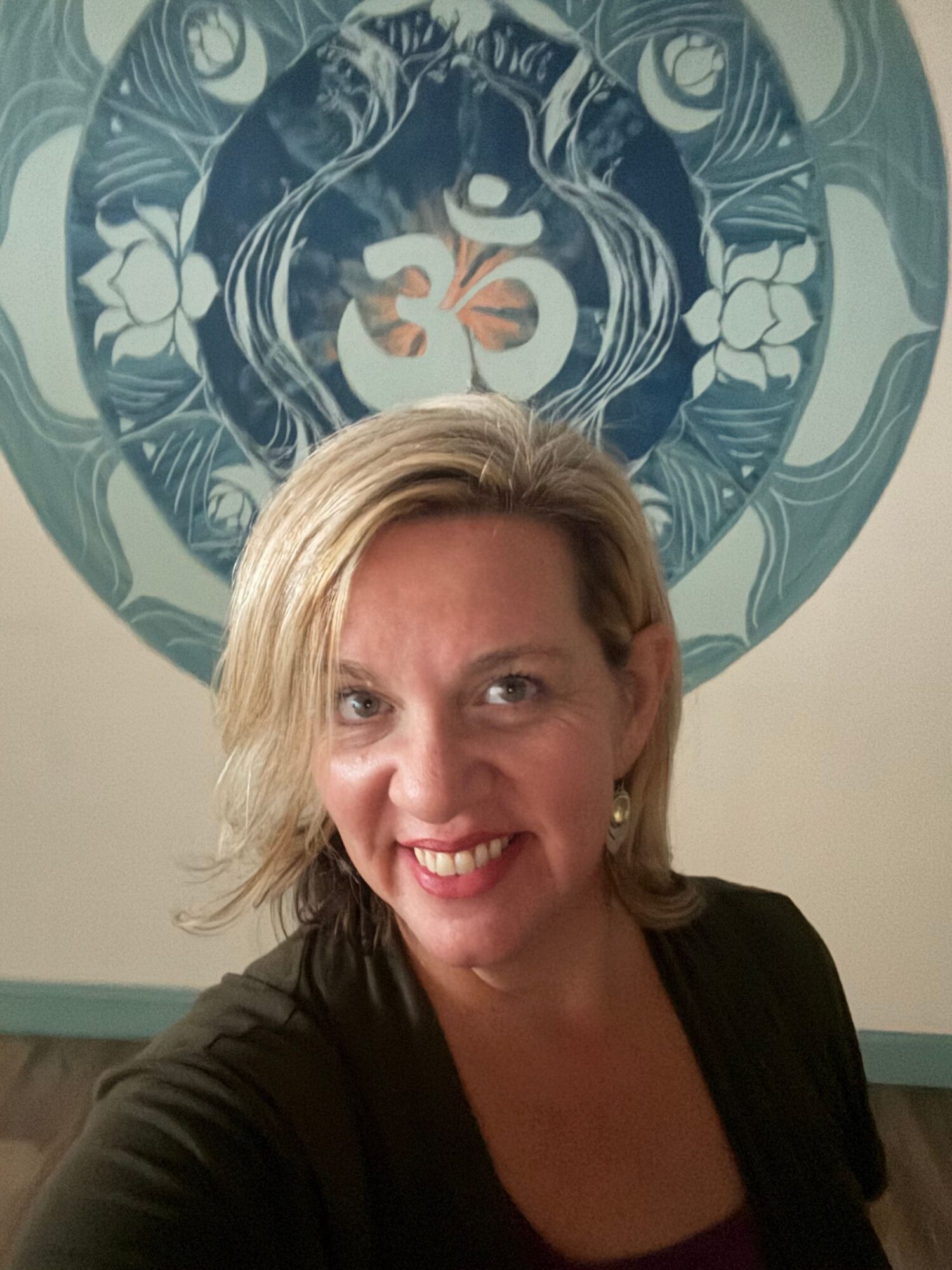 Woman with blonde hair smiling, taking a selfie in front of a decorative circular wall art.