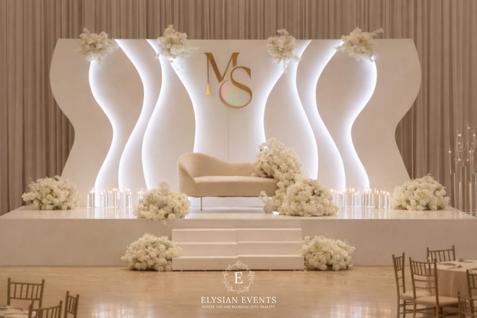 Elegant event stage with white floral decorations, a white sofa, and a backdrop with gold initials 'MS' and 'Elysian Events'.