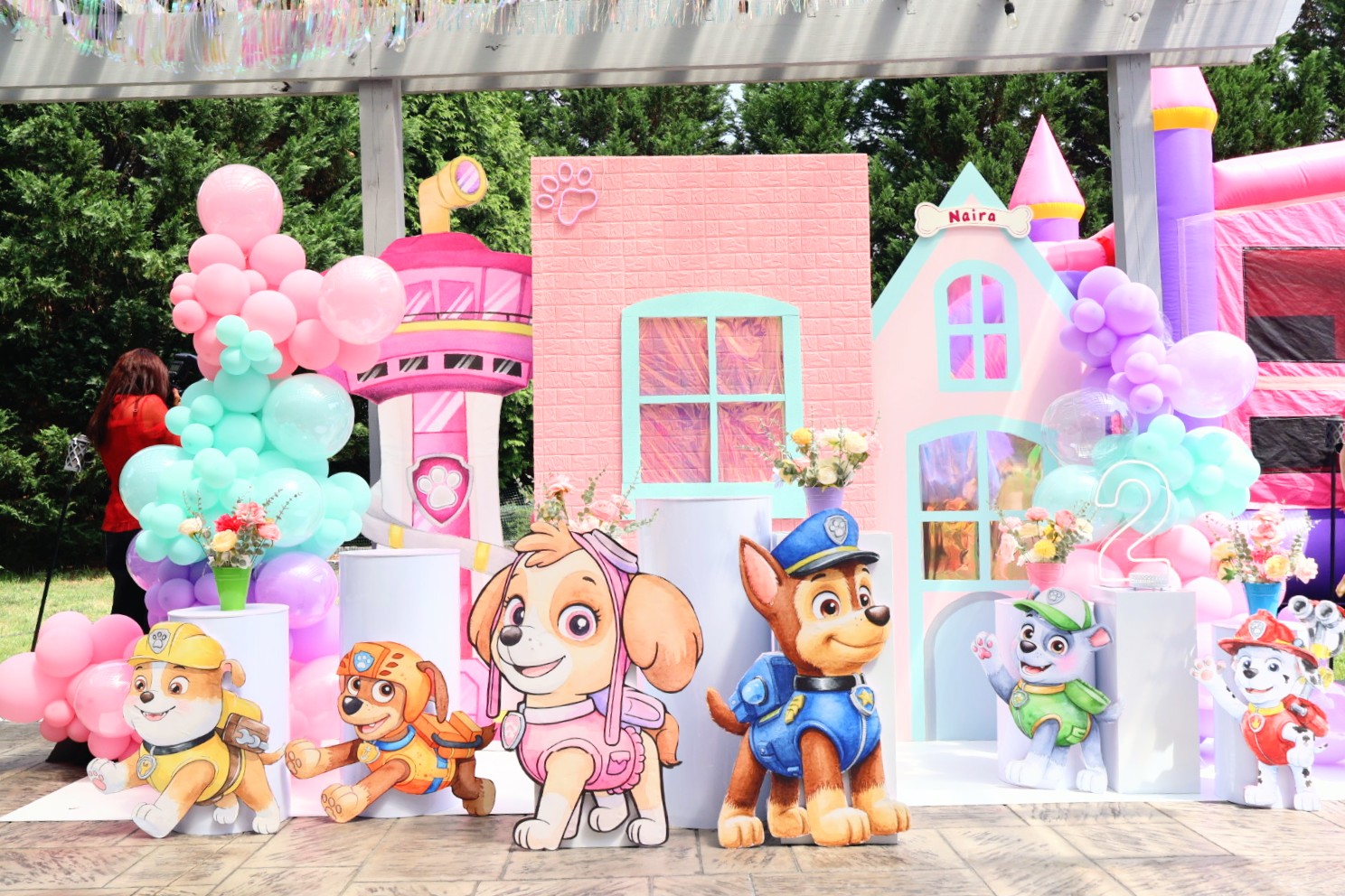 Colorful birthday party setup with balloons, cartoon characters, and a pink castle backdrop, outdoors with trees in the background.