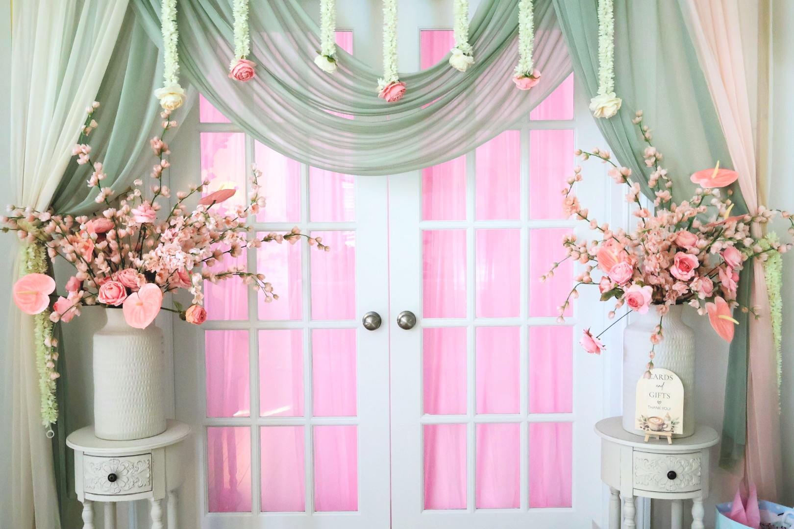 Double door with pink curtains, floral arrangements on white vases, green drapes, and pink lighting, decorated for an event.