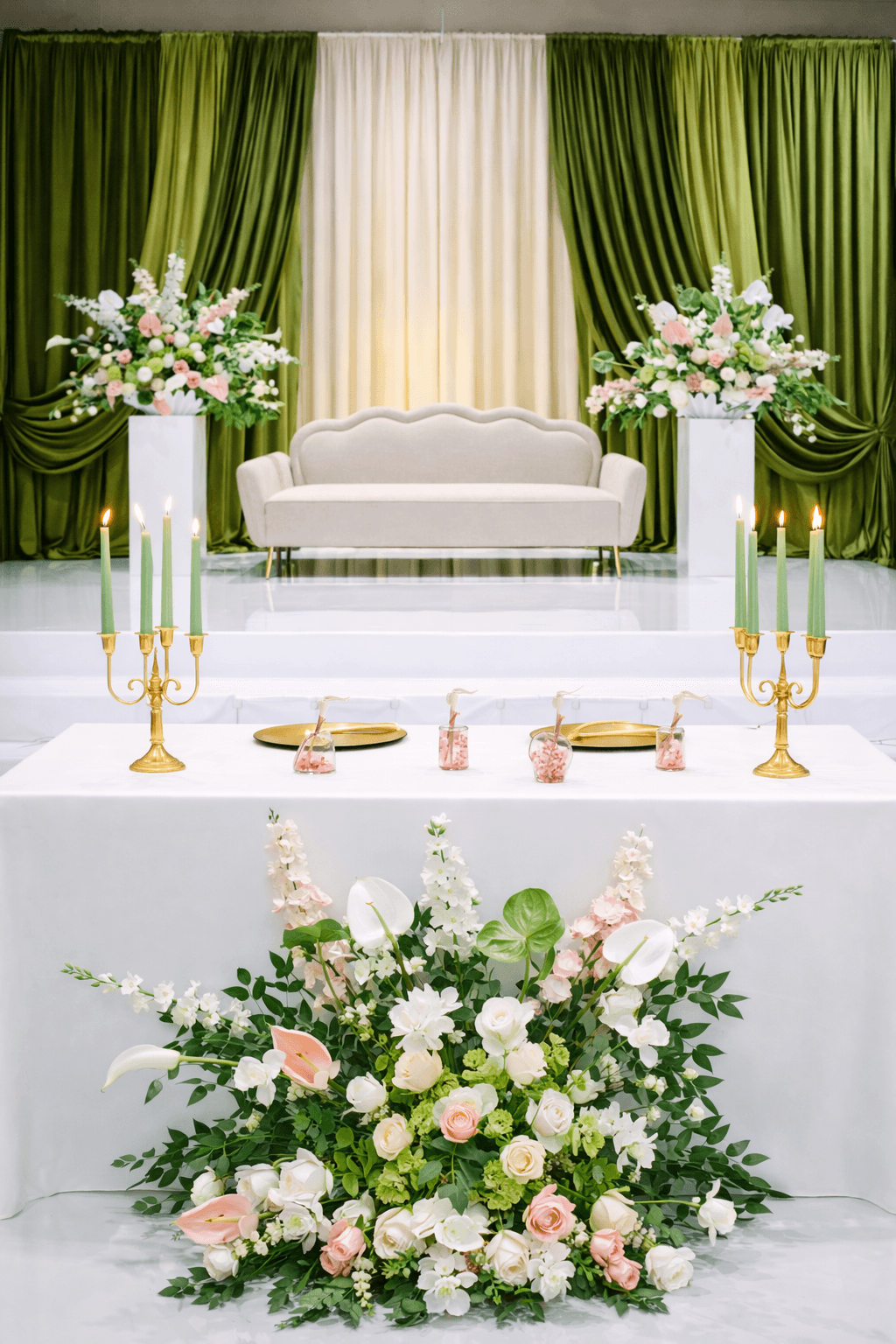 Elegant room with green curtains, white sofa, floral arrangements, and a decorated table, possibly for a celebration or event.