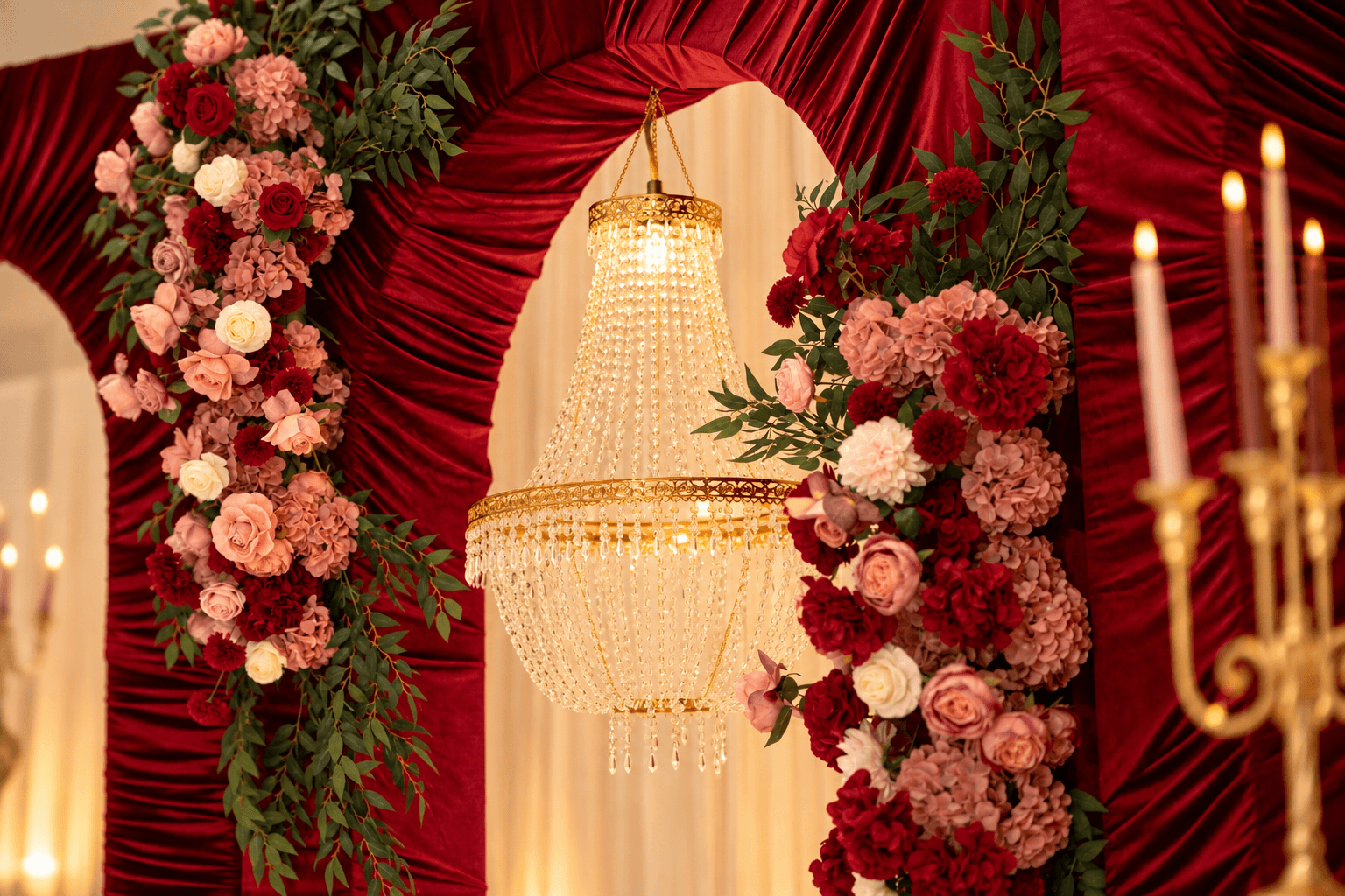 Elegant chandelier hanging in front of red draped curtains with floral arrangements and candelabra, festive setting.
