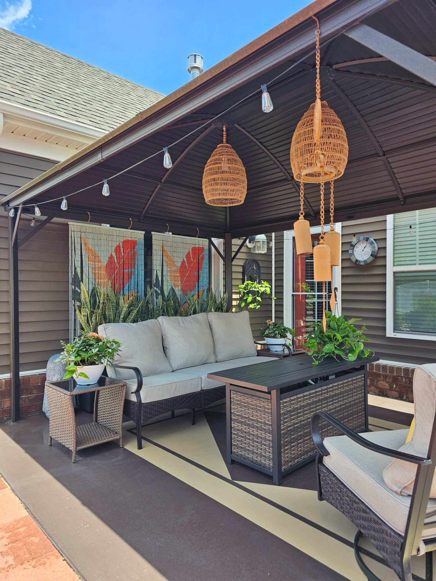 Outdoor patio with a beige sofa, wicker chairs, a black table, and hanging wicker pendant lights under a covered roof.