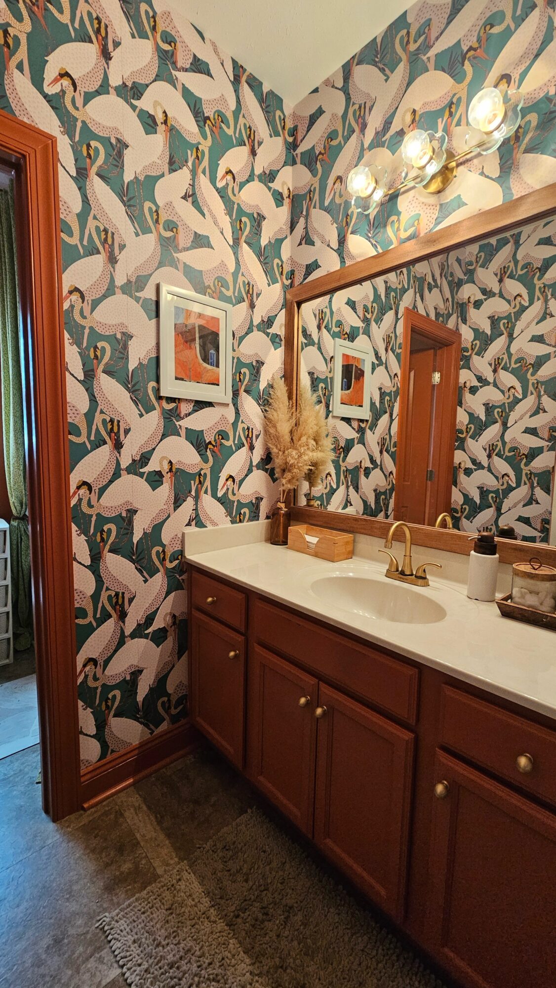 Bathroom vanity with a mirror, floral wallpaper, and a sink with a gold faucet, wooden cabinets, and framed pictures on the wall.