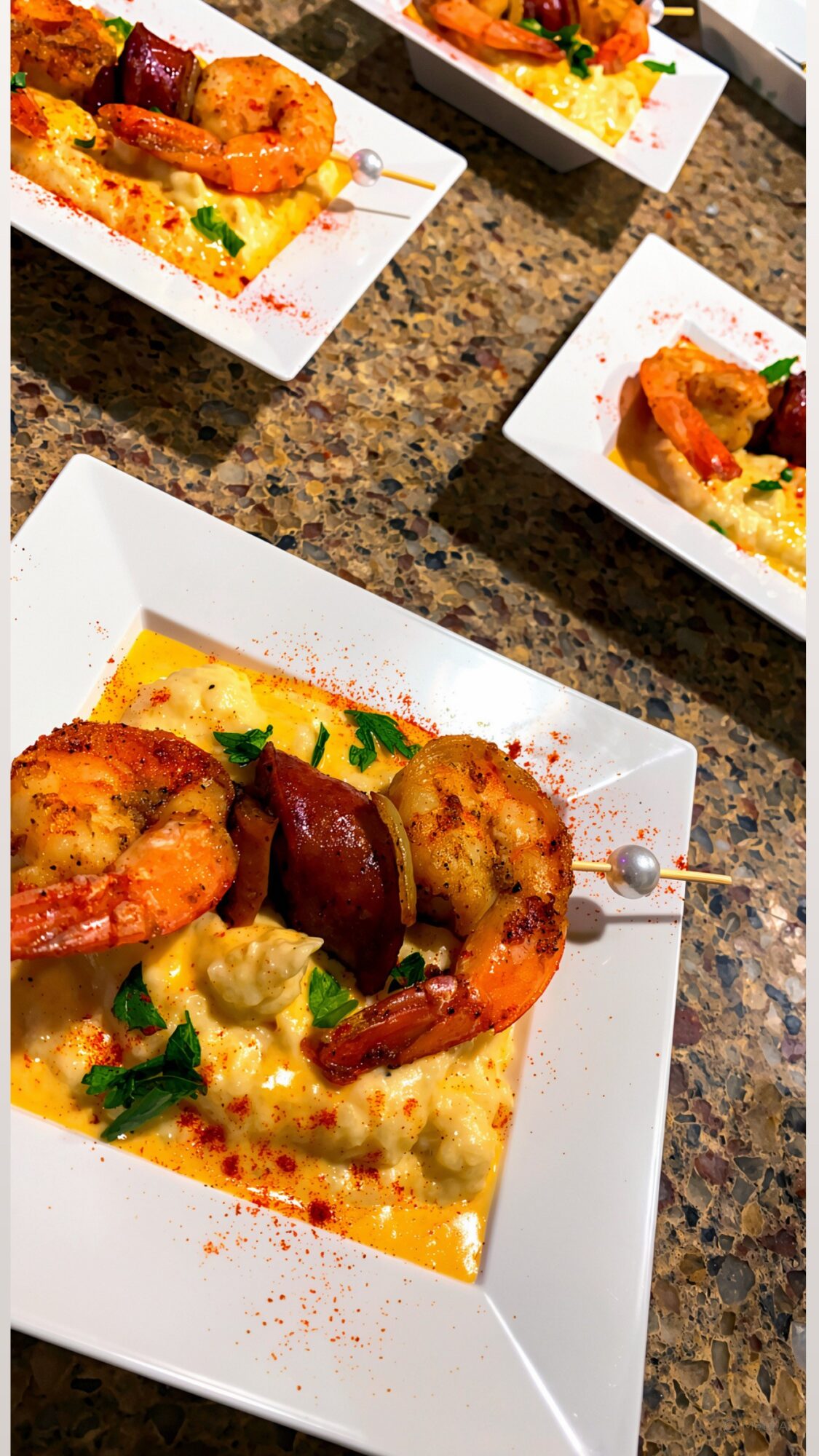 Close-up of a white square plate with skewered grilled shrimp and sausage on a bed of creamy sauce, garnished with herbs.