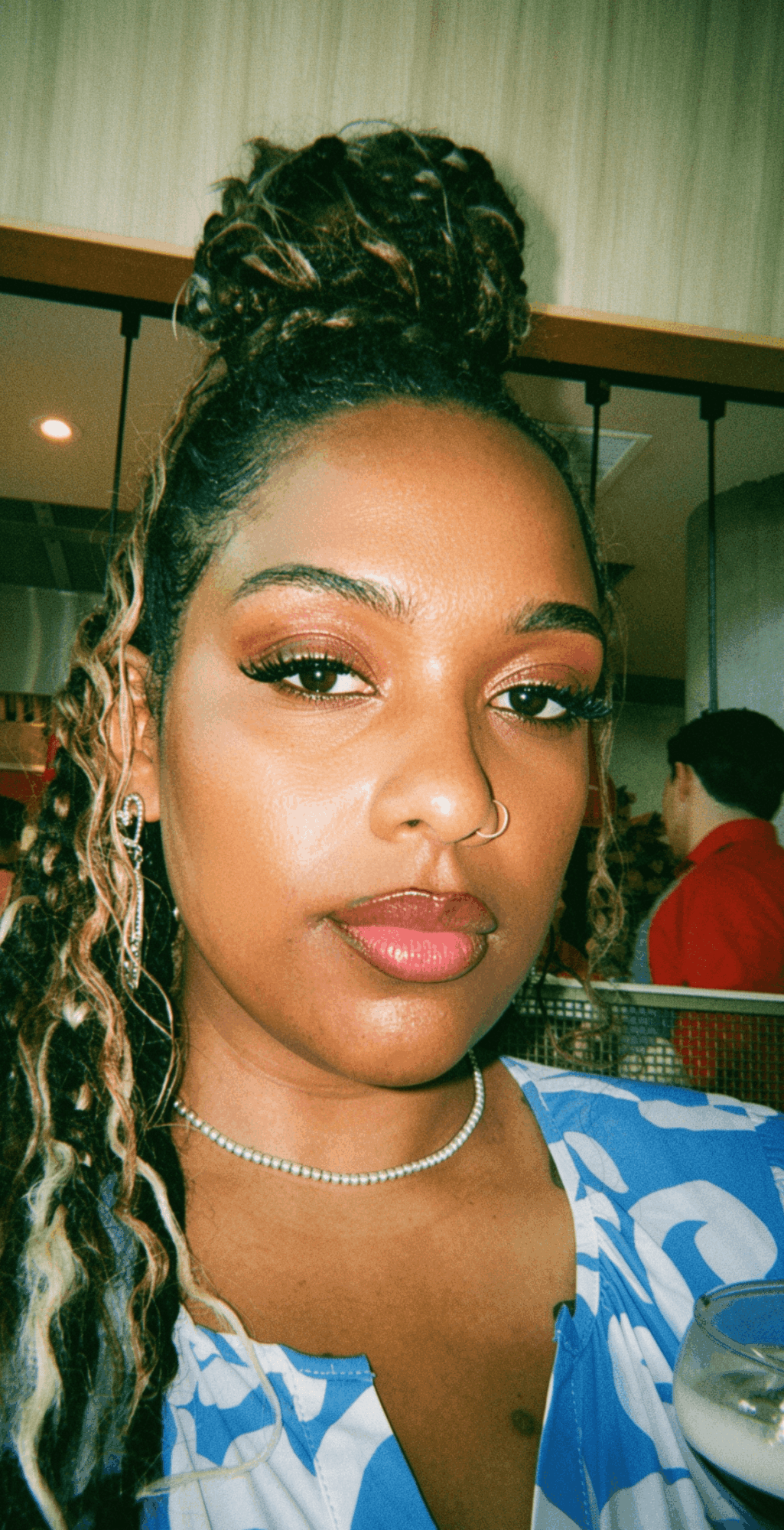 Young woman with braided hair styled in an updo, wearing makeup, a necklace, and a blue patterned top, indoors.