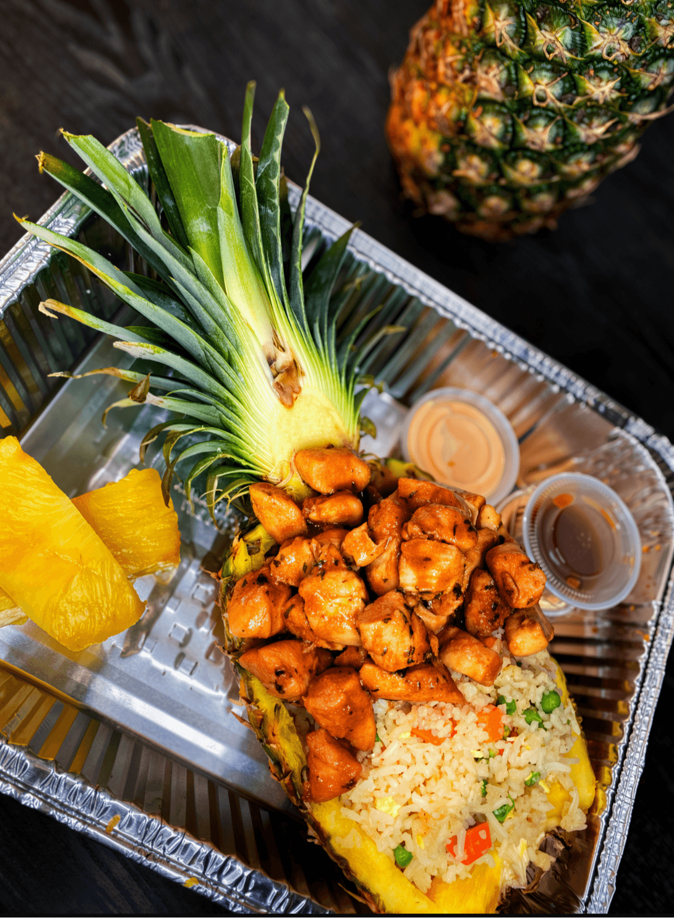 Pineapple with chopped pineapple, rice, and sauces in a tray, with whole pineapple in background.