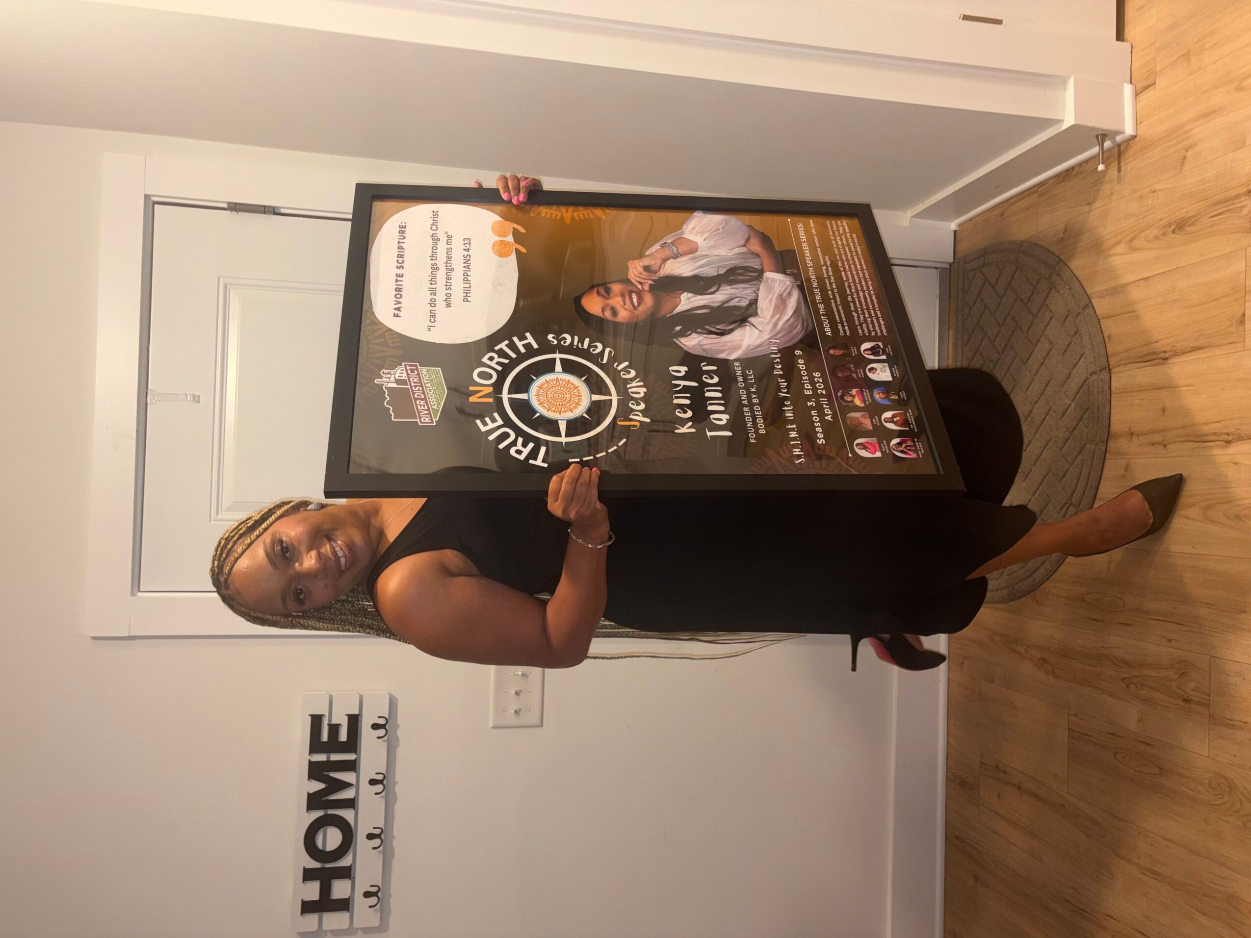 Woman in black dress holding a large framed award or poster, standing indoors near a white door and wall with a 'HOME' sign, wooden floor, and a round black rug.