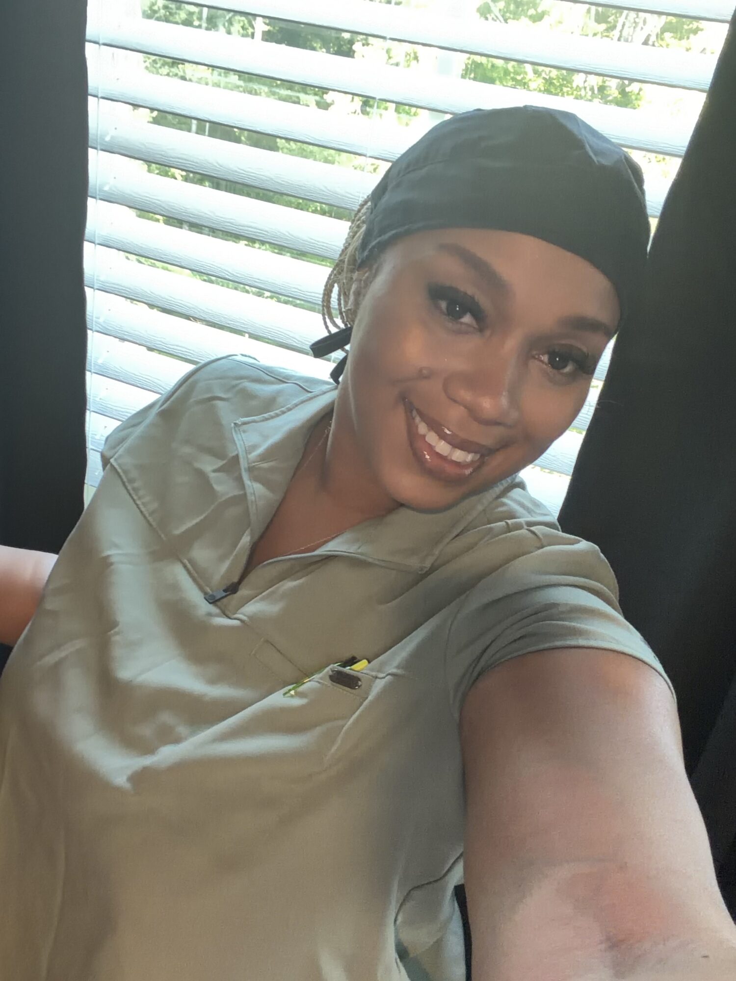 Smiling woman wearing a headscarf and light-colored top, taking a selfie indoors with window blinds behind her.