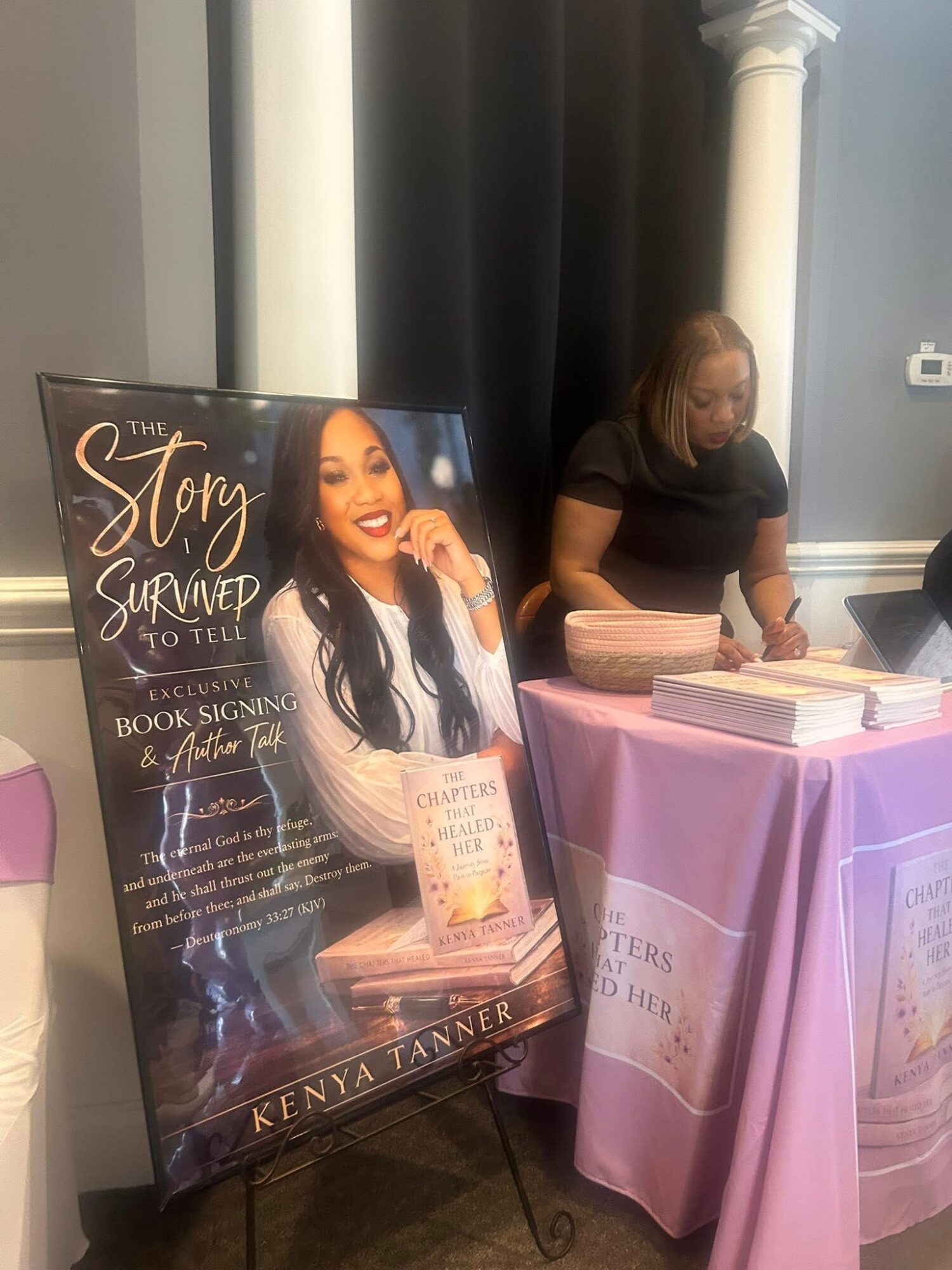 Woman at a table with books and a sign promoting her story and book signing event.
