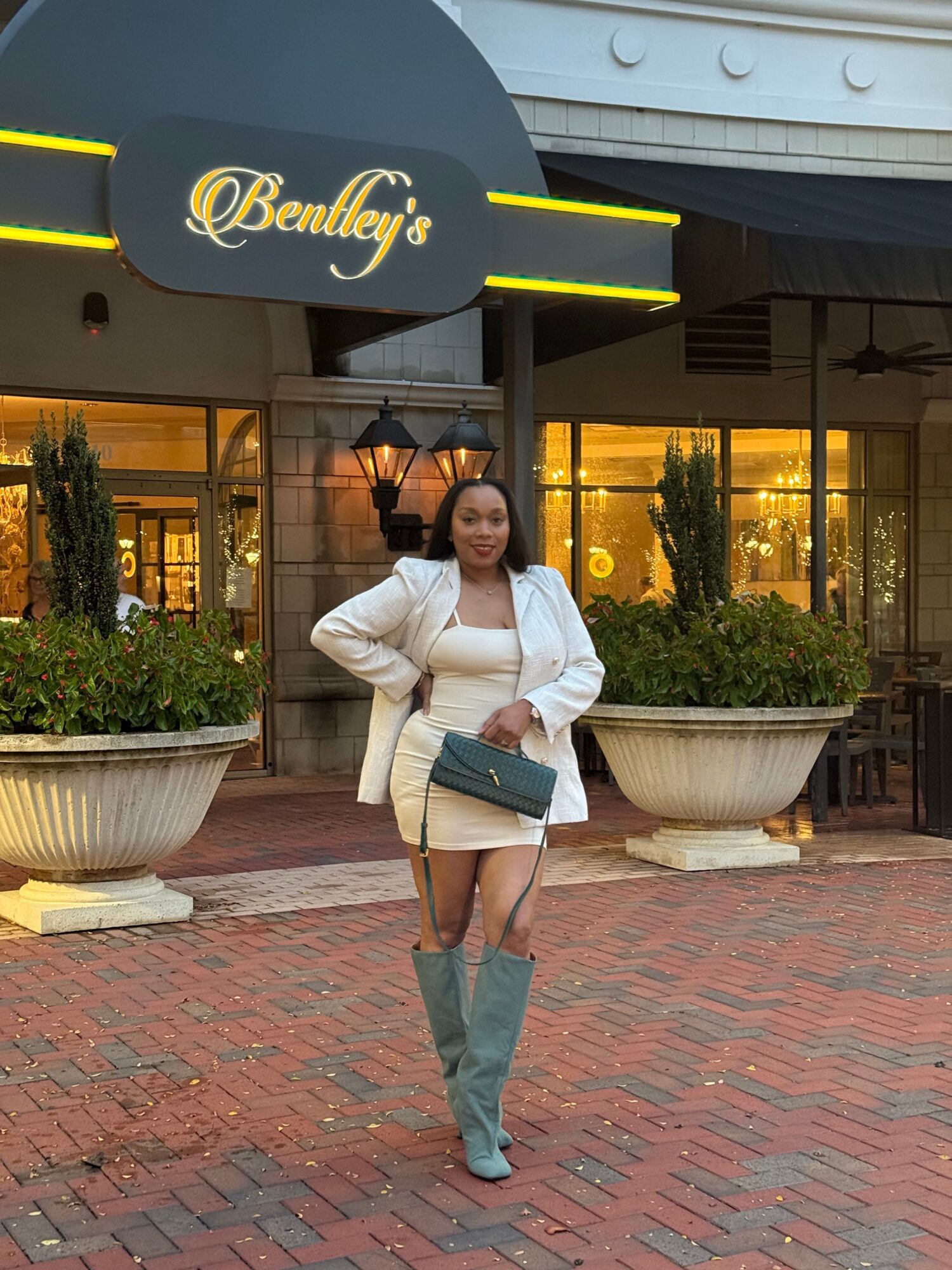 Woman standing on brick sidewalk outside building holding a laptop, wearing a white blazer and gray pants.