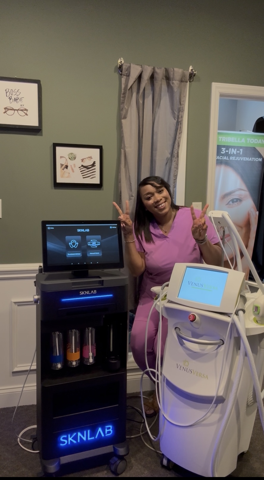 Woman in pink scrubs smiling and giving a thumbs-up in a medical room with equipment and a green wall.
