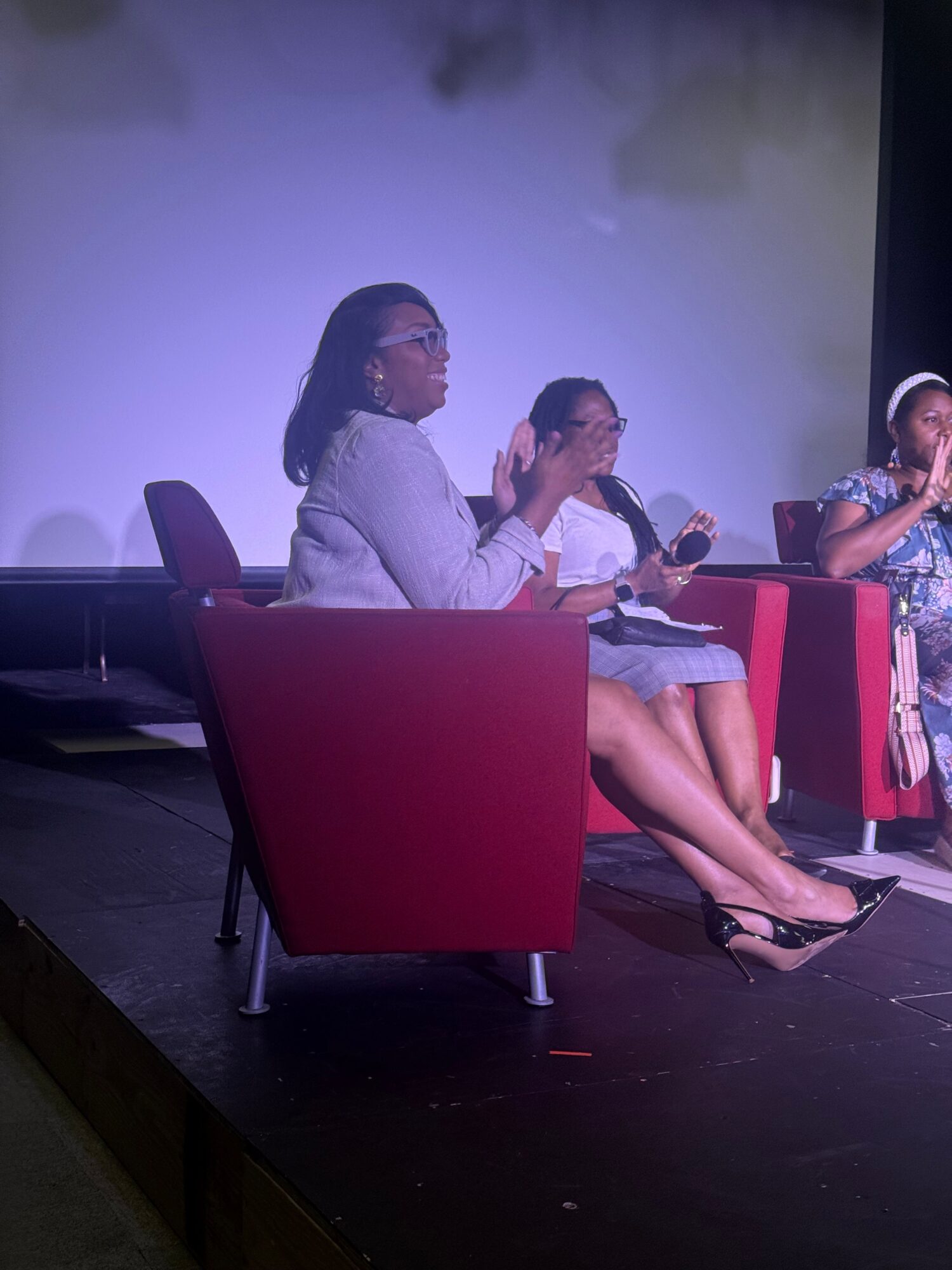 Two women sitting on red chairs, one holding a microphone, in a room with a plain background.