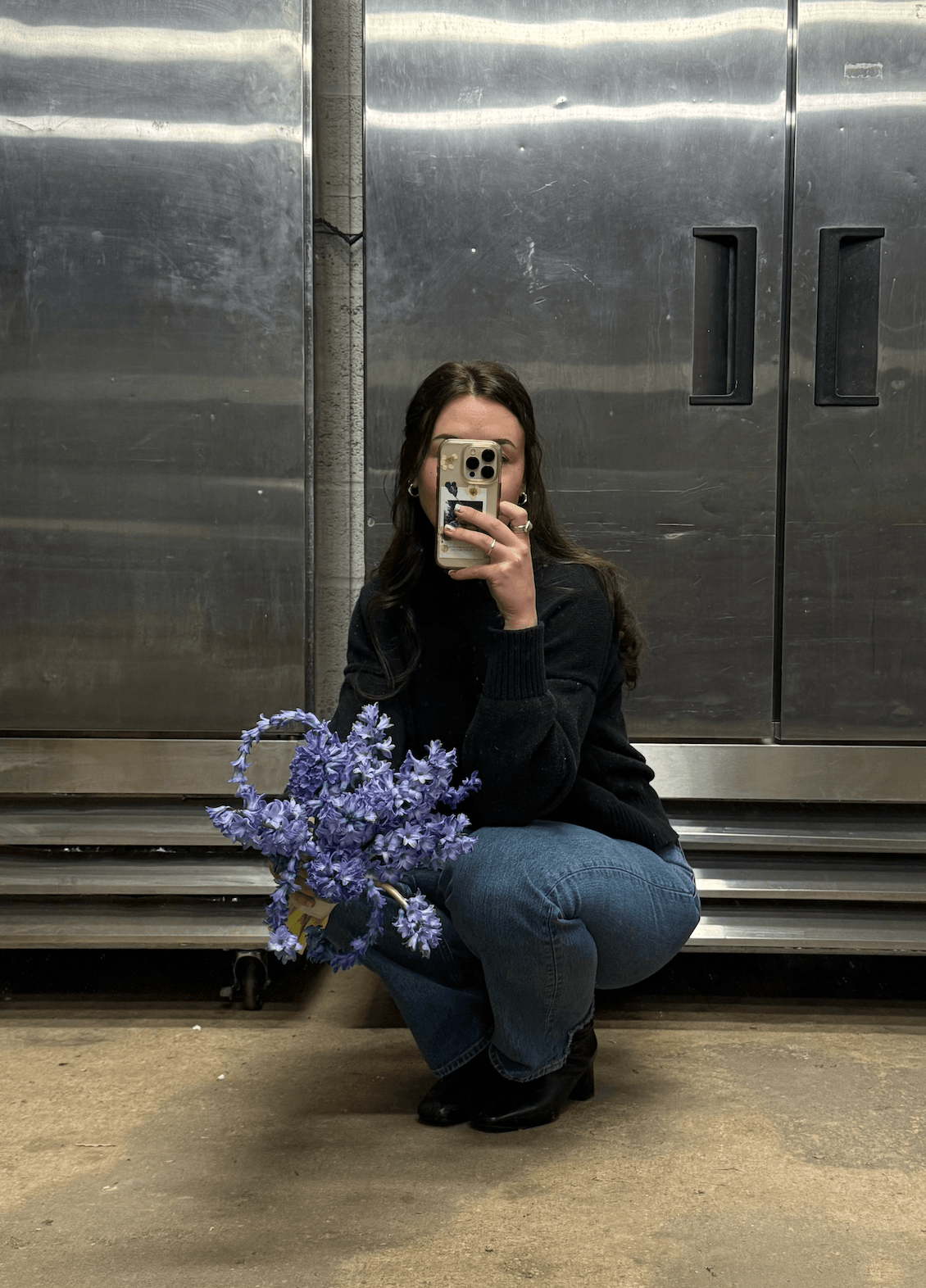 Person crouching with a bouquet of purple flowers, taking a mirror selfie in an industrial setting, metal doors in background.