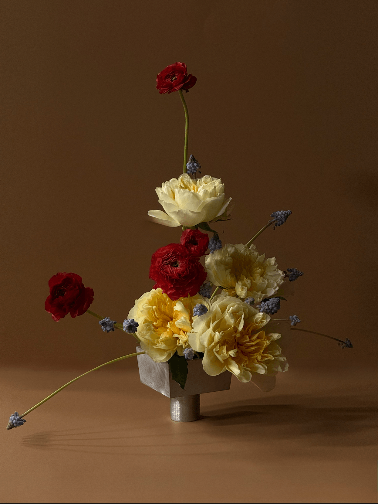 A flower arrangement with red, yellow, and white flowers in a square vase on a wooden surface, against a brown background.