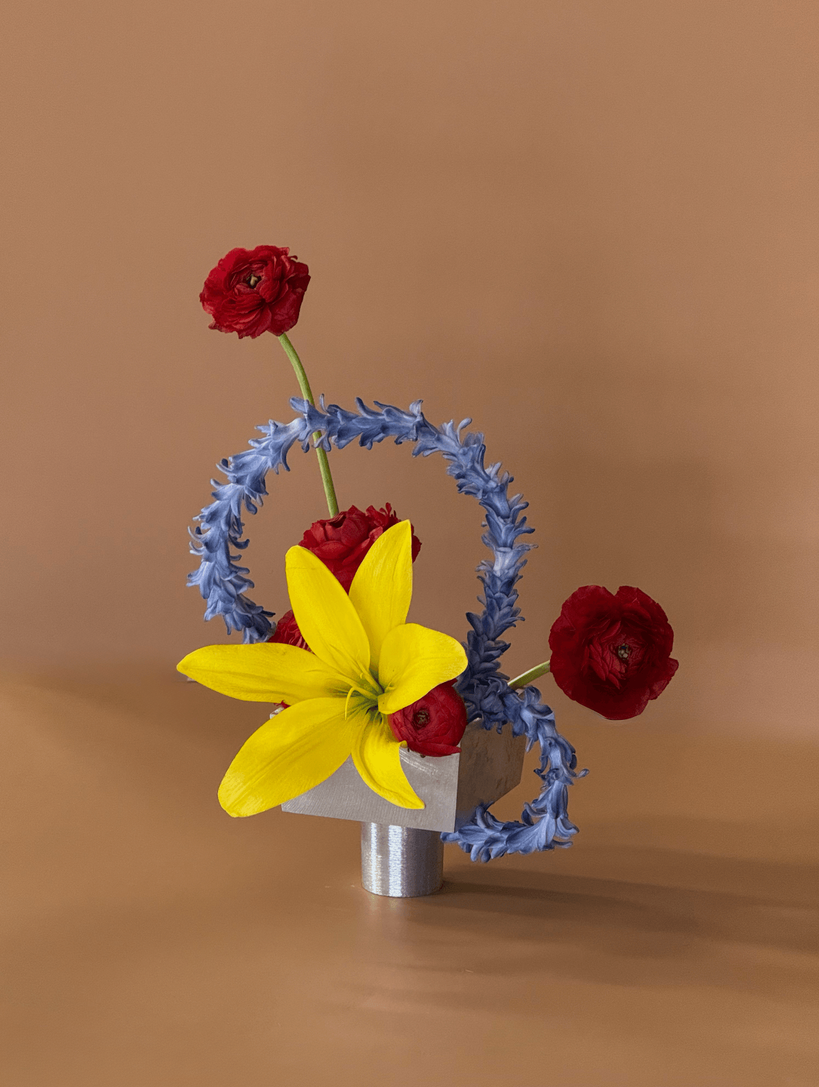 Arrangement of red, yellow, and blue flowers in a small metallic vase against a plain background.